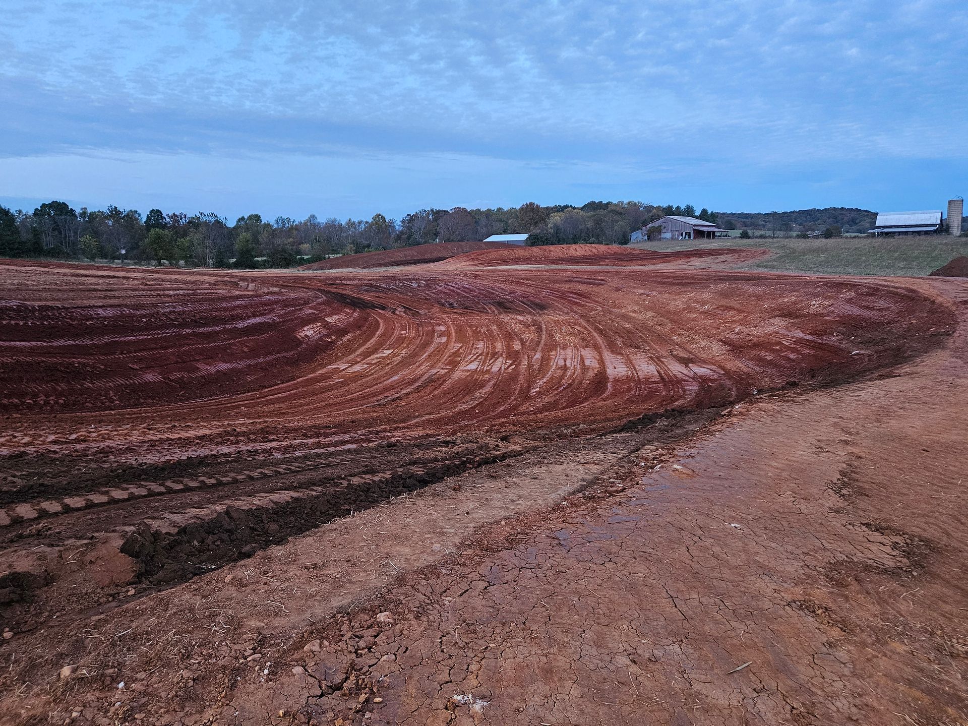 Freshly graded red dirt field under a blue sky, with trees and buildings in the distance