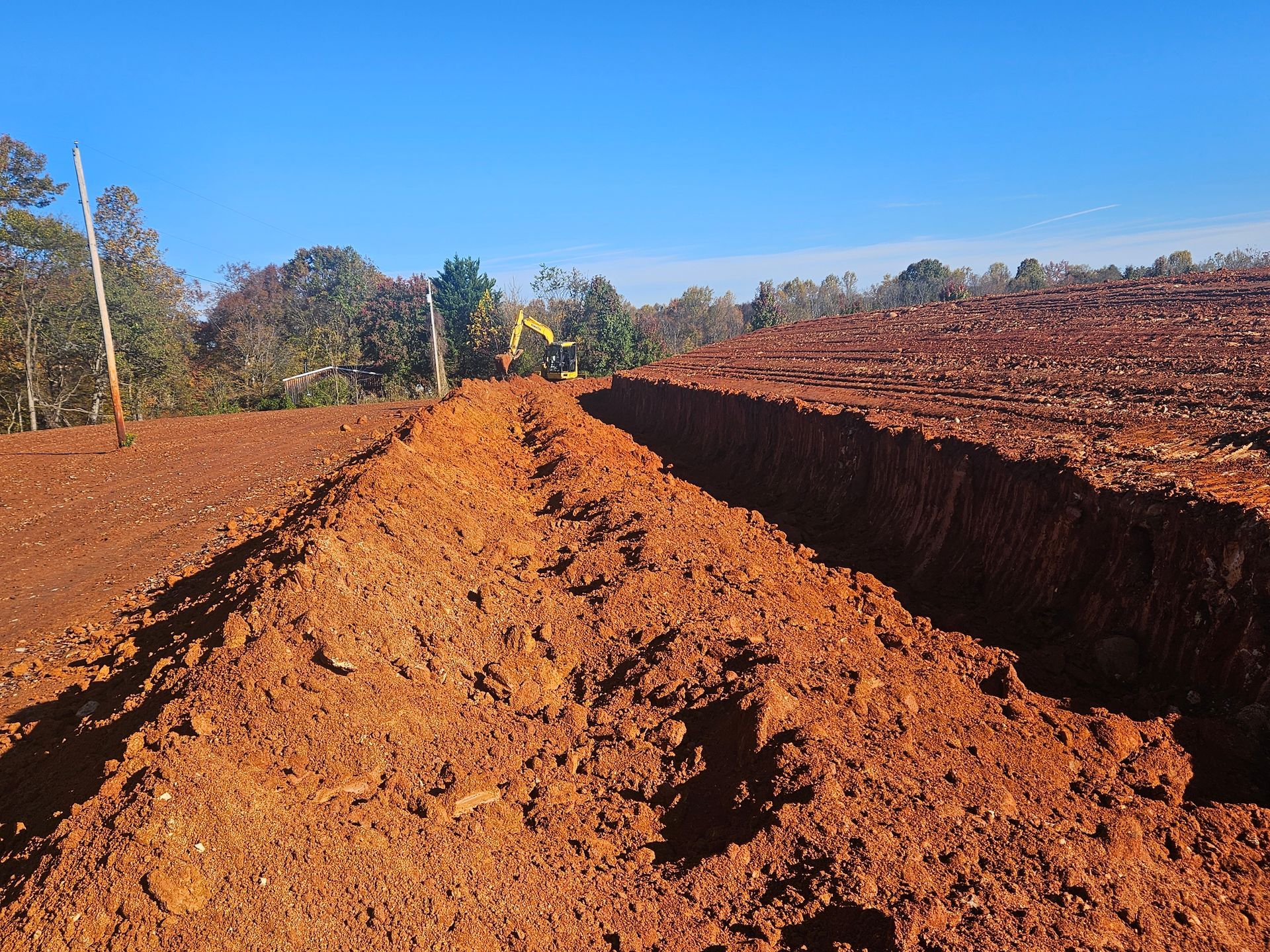 Red dirt road cut through a muddy hillside under a clear blue sky, with trees in the distance.