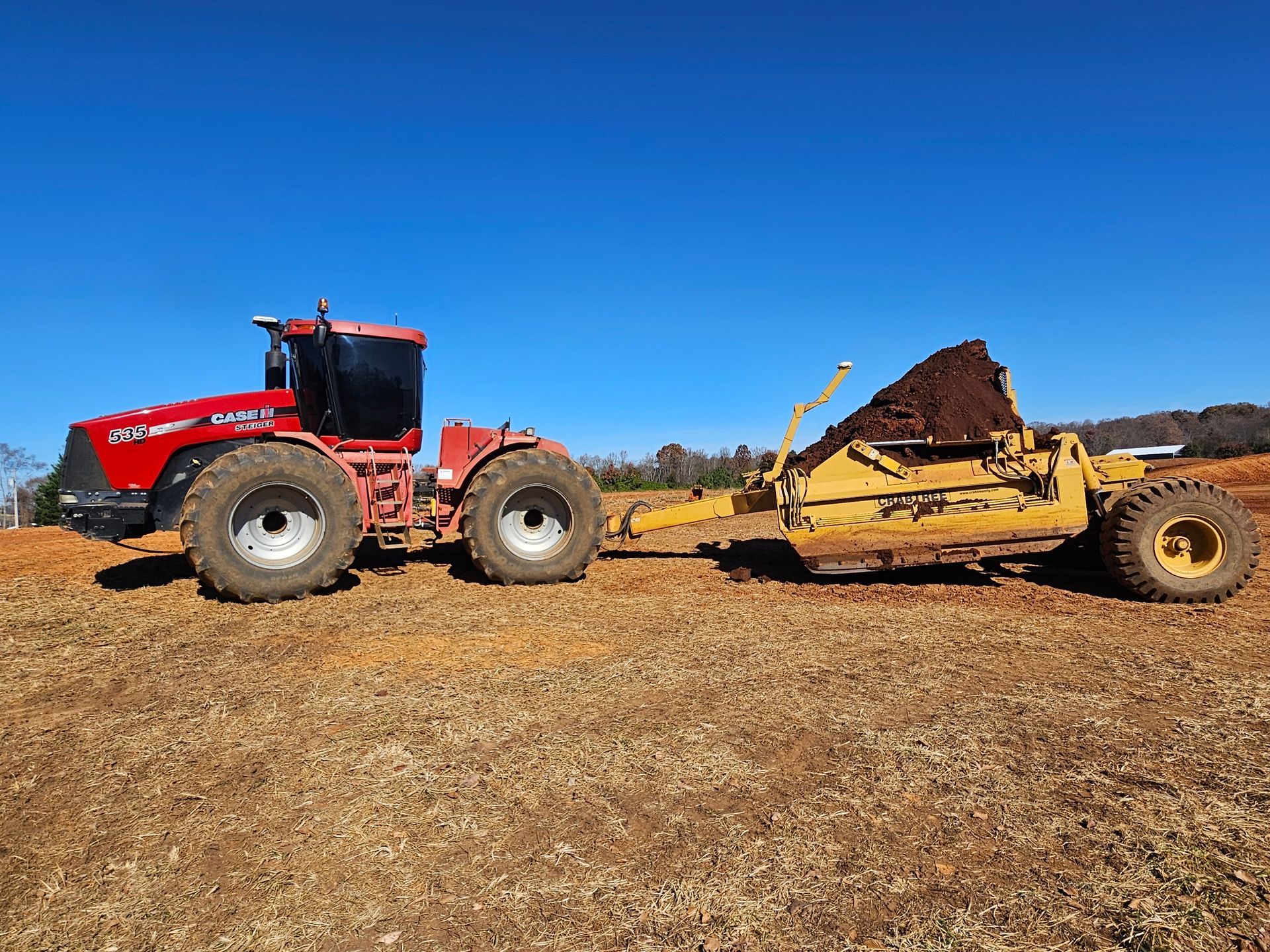Red tractor pulling a yellow manure spreader loaded with dirt across a field under a blue sky