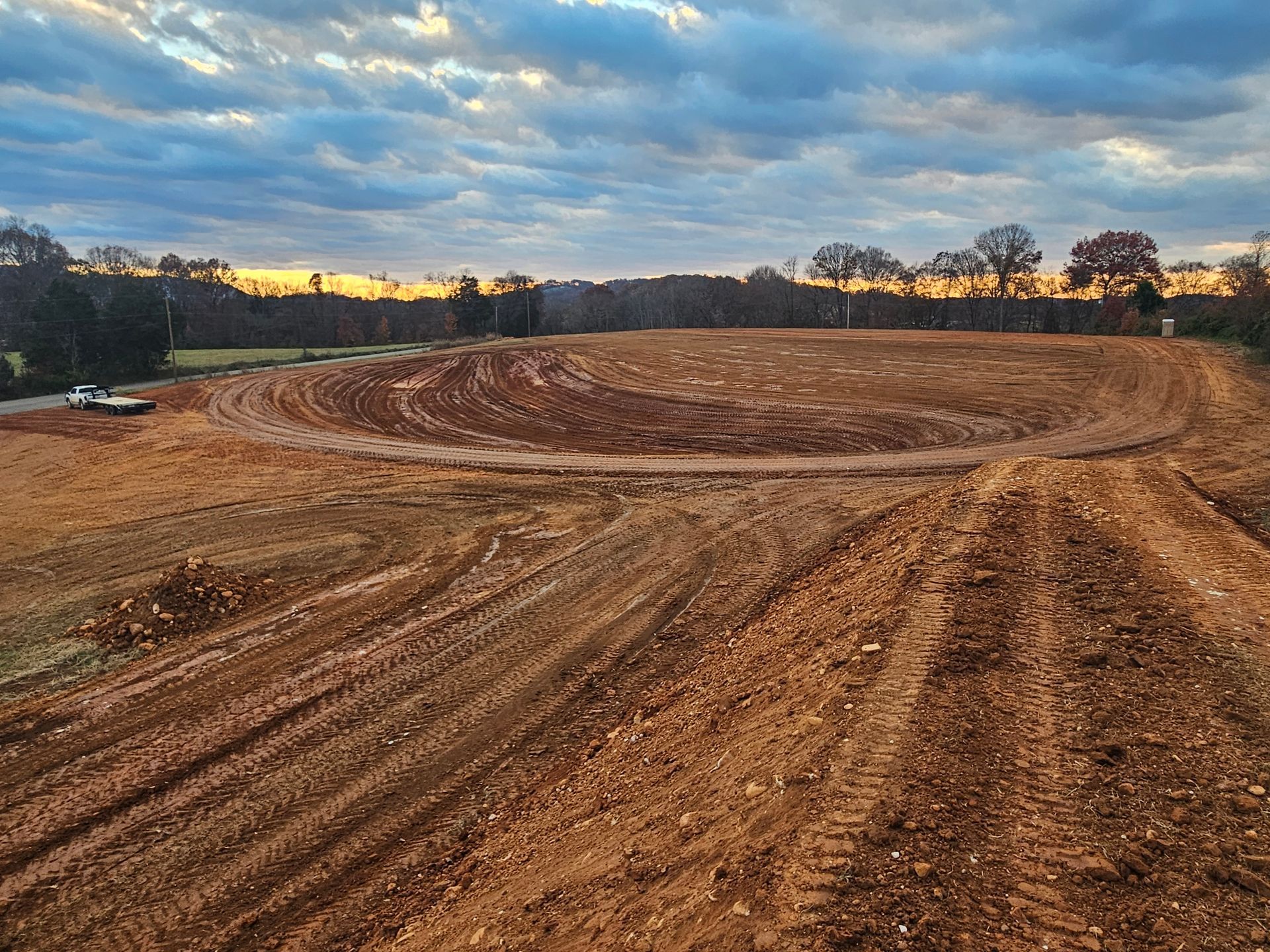 Freshly plowed dirt field with curved tire tracks under a cloudy sunset sky