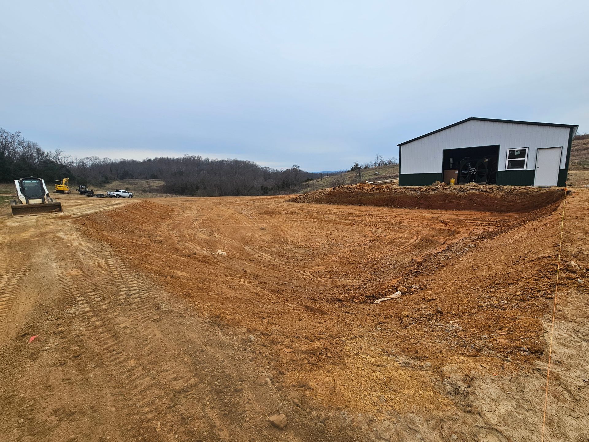 Dirt lot with a white building on the right and a tractor in the distance under an overcast sky