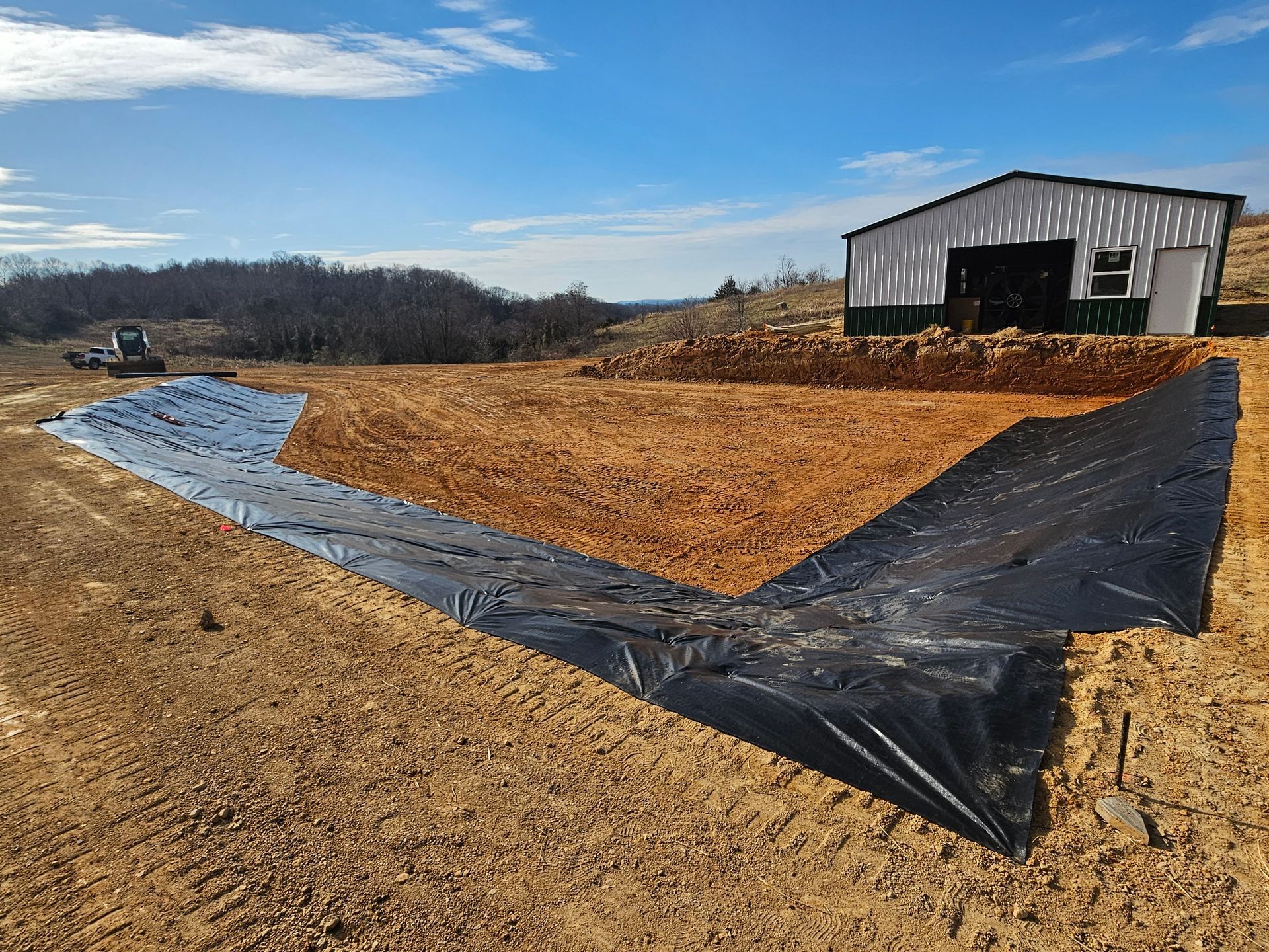 Black plastic-lined site with tan dirt fill beside a white metal shed under a blue sky.