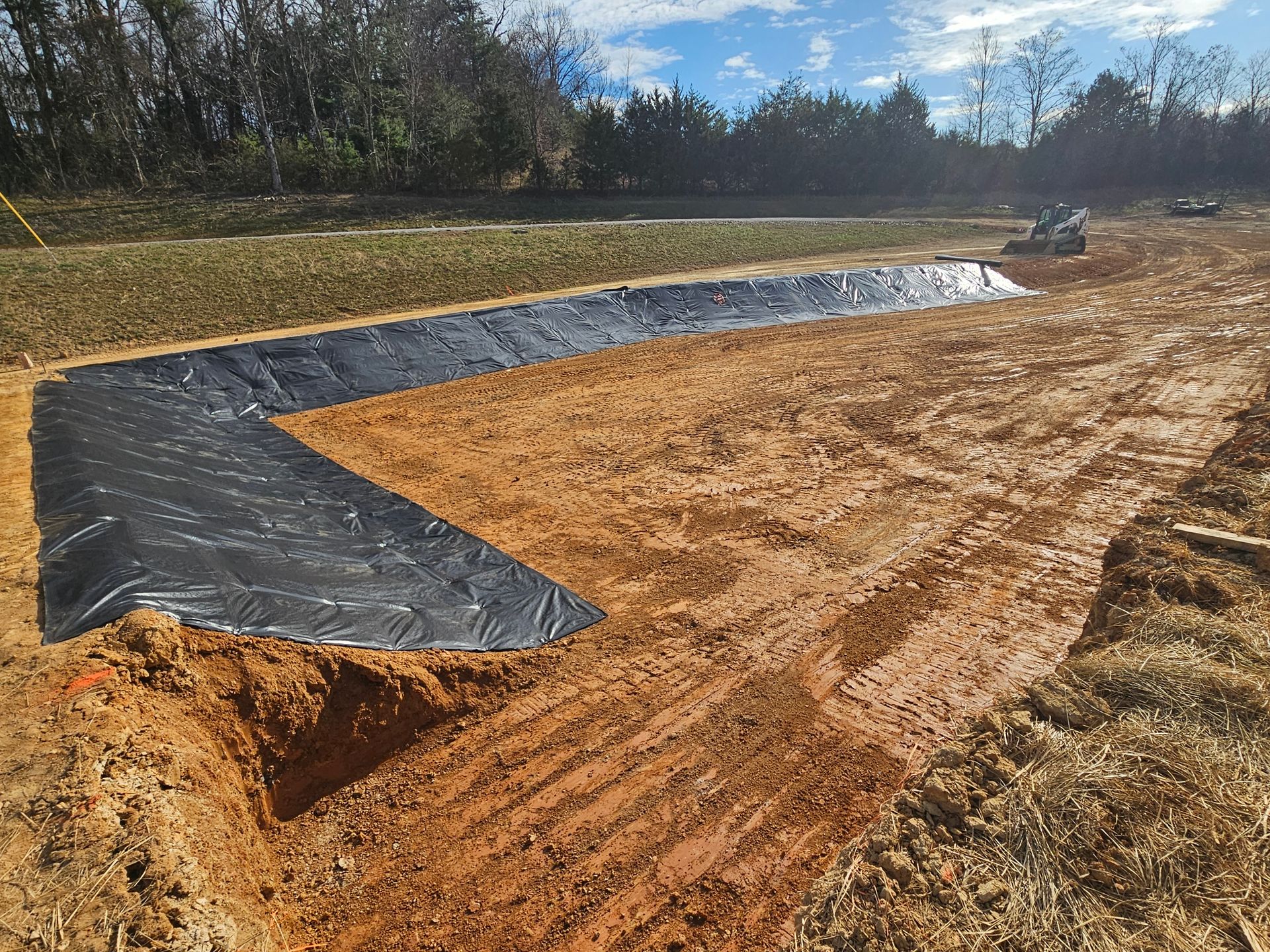 Black plastic liner in a newly dug retention pond with exposed dirt and grassy edges under a clear sky