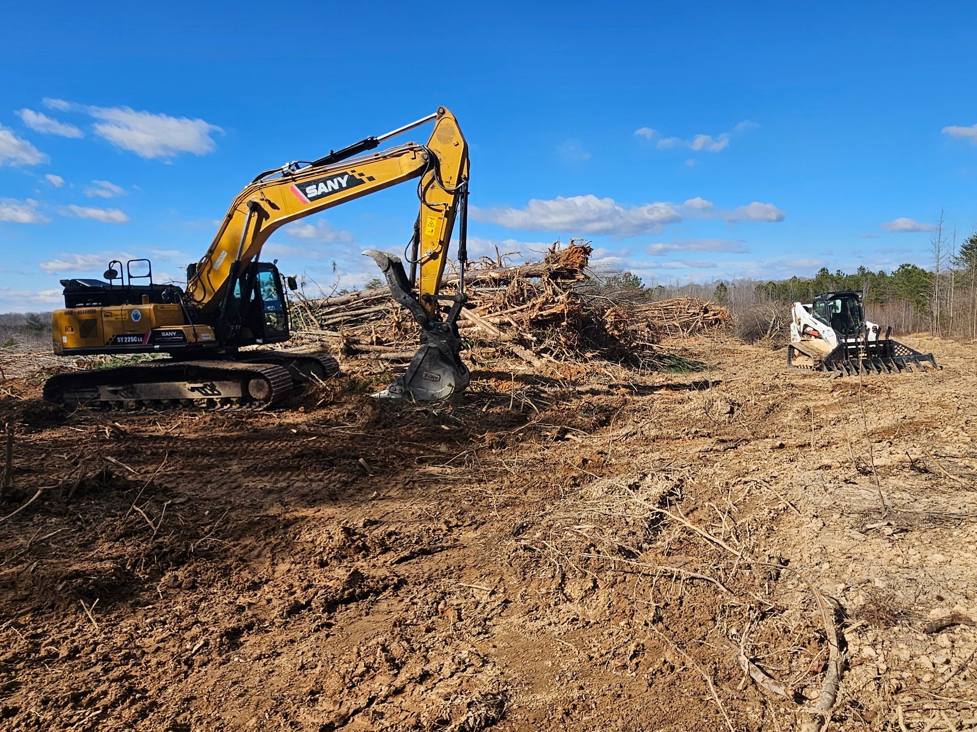 Yellow excavator clearing a muddy construction site with piles of debris under a blue sky