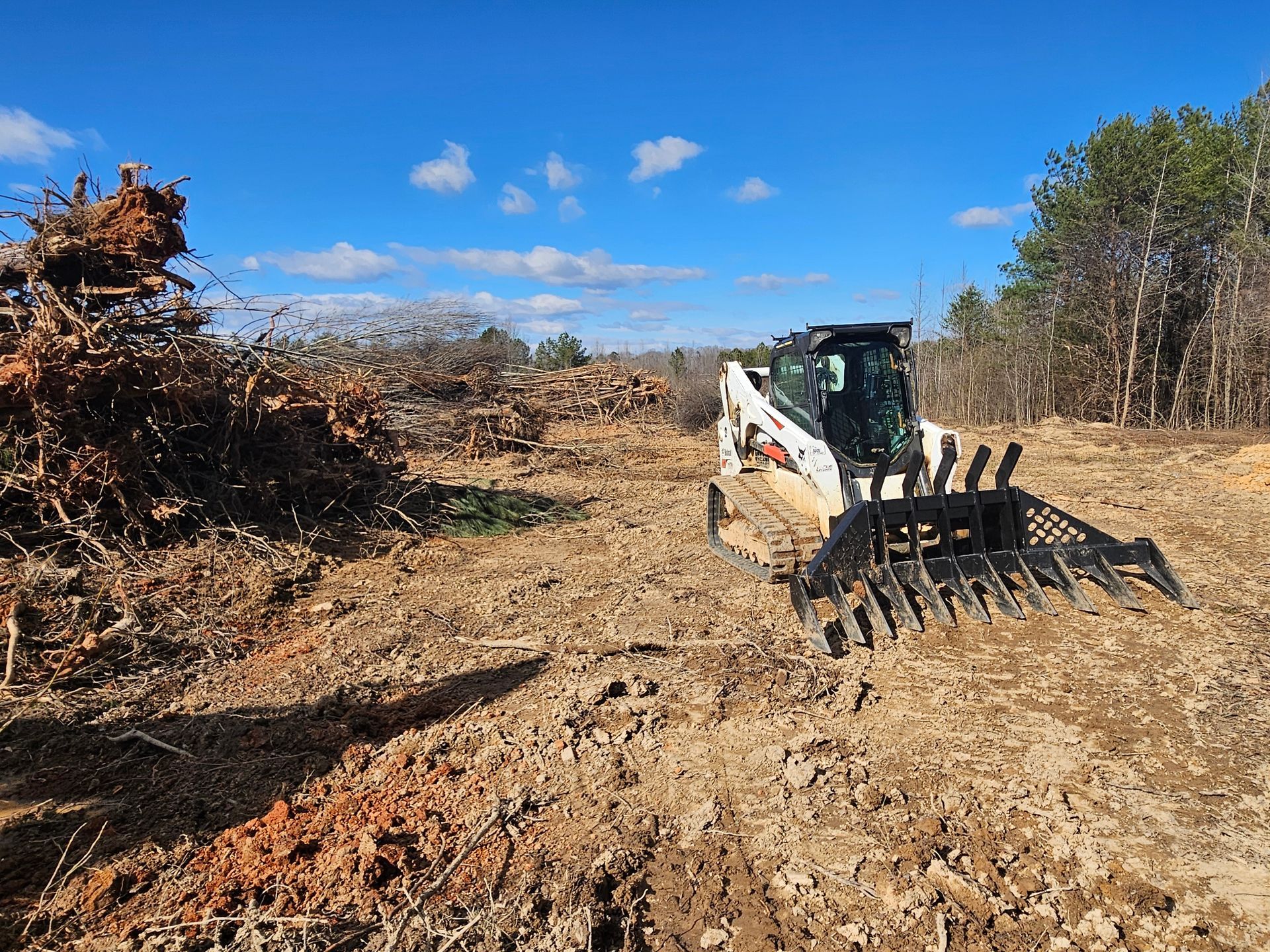 White skid steer with grapple bucket clearing brush in a dirt field under a blue sky