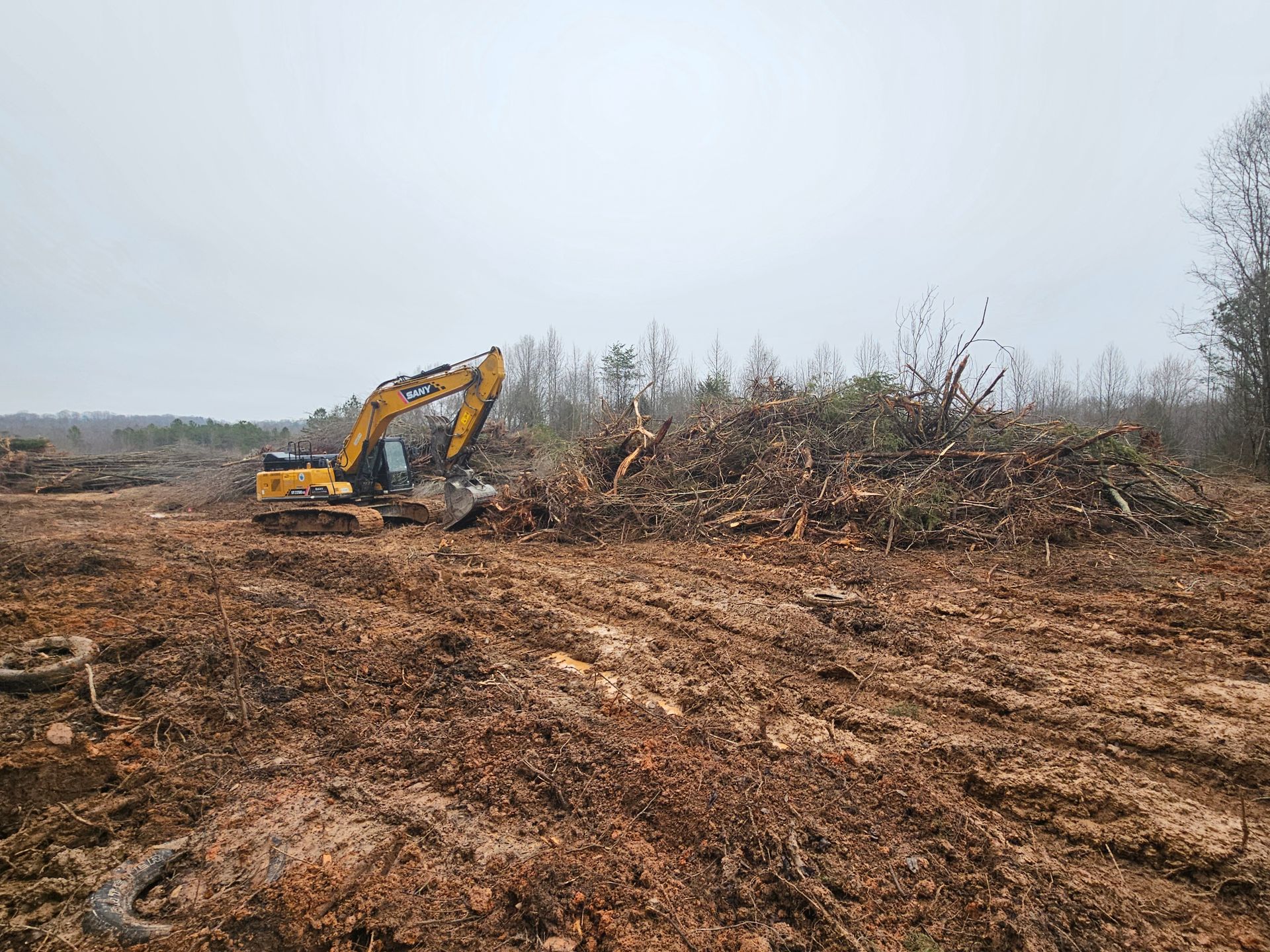 Yellow excavator clearing a muddy, brush-covered field under a gray sky