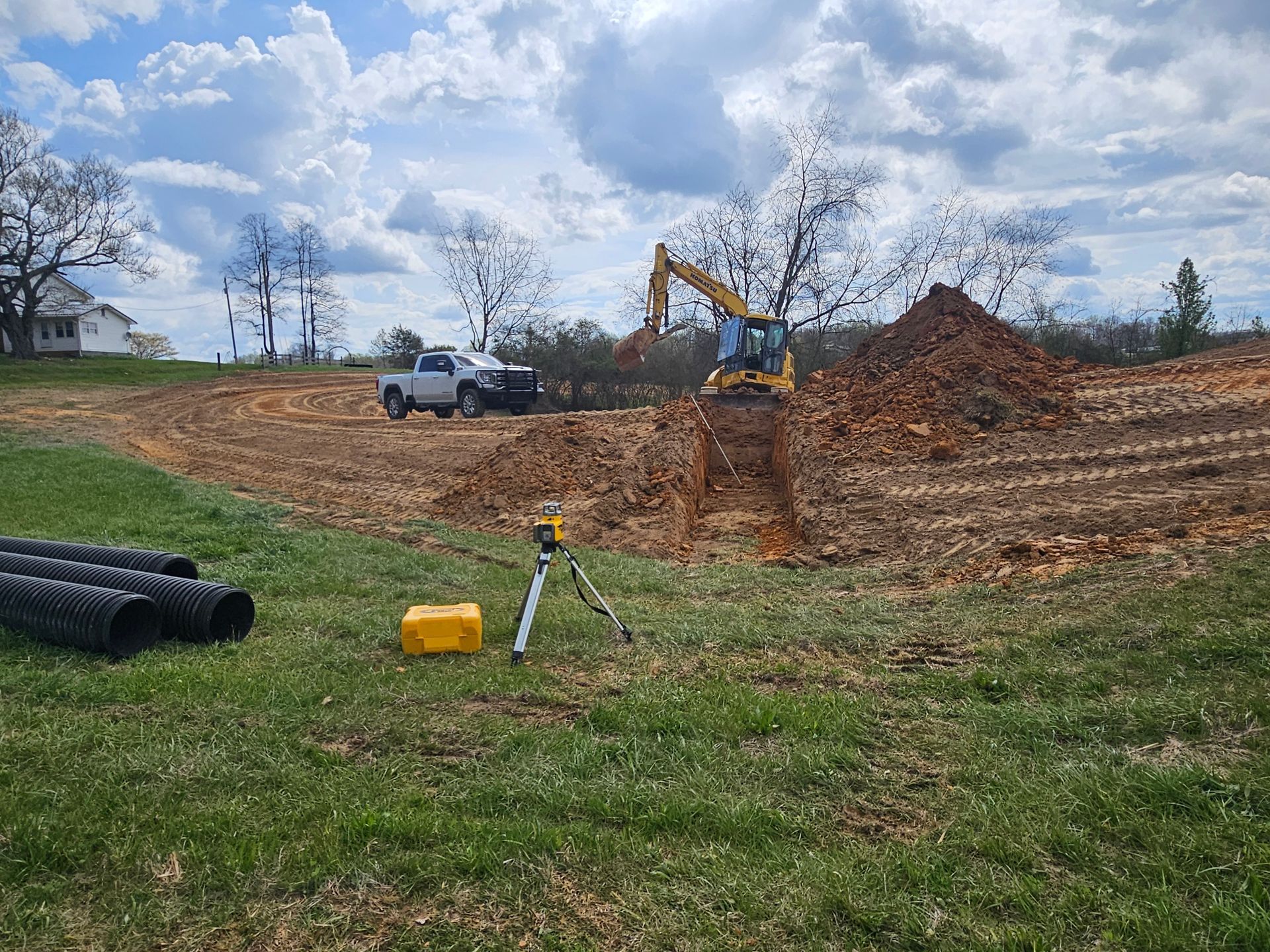 Construction excavator clearing dirt on a rural hillside with a white truck and survey tripod in the foreground