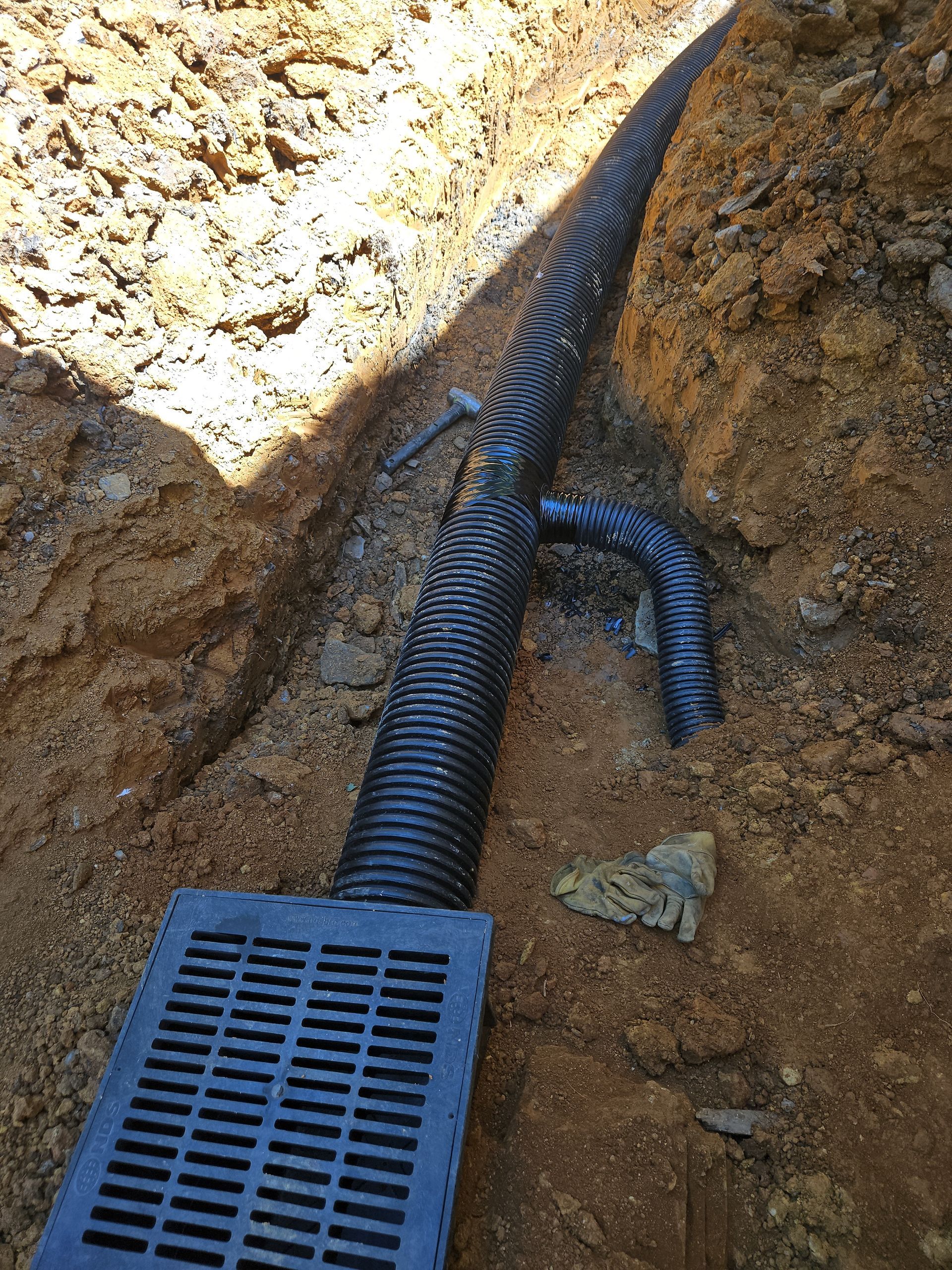 Black drainage pipe in a dirt trench connected to a grating drain inlet.
