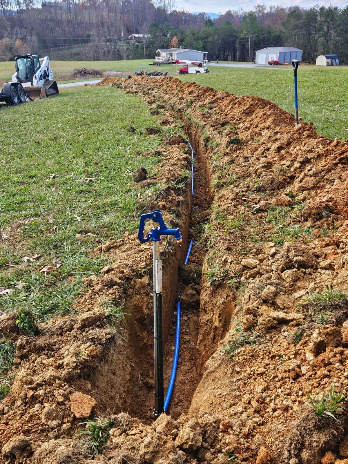 Trench with blue irrigation pipe and sprinkler head running through a grassy field near houses