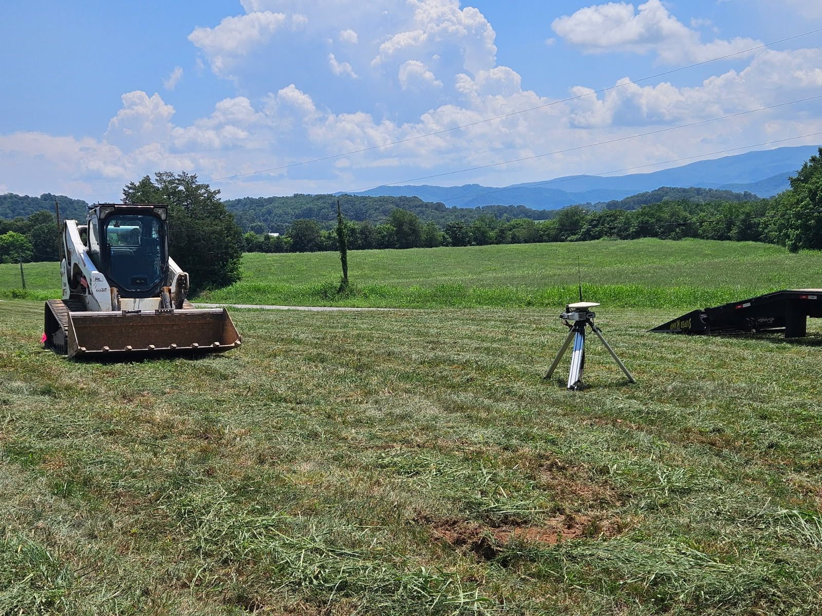 Skid steer mower cutting a grassy field under a blue sky with mountains in the distance.