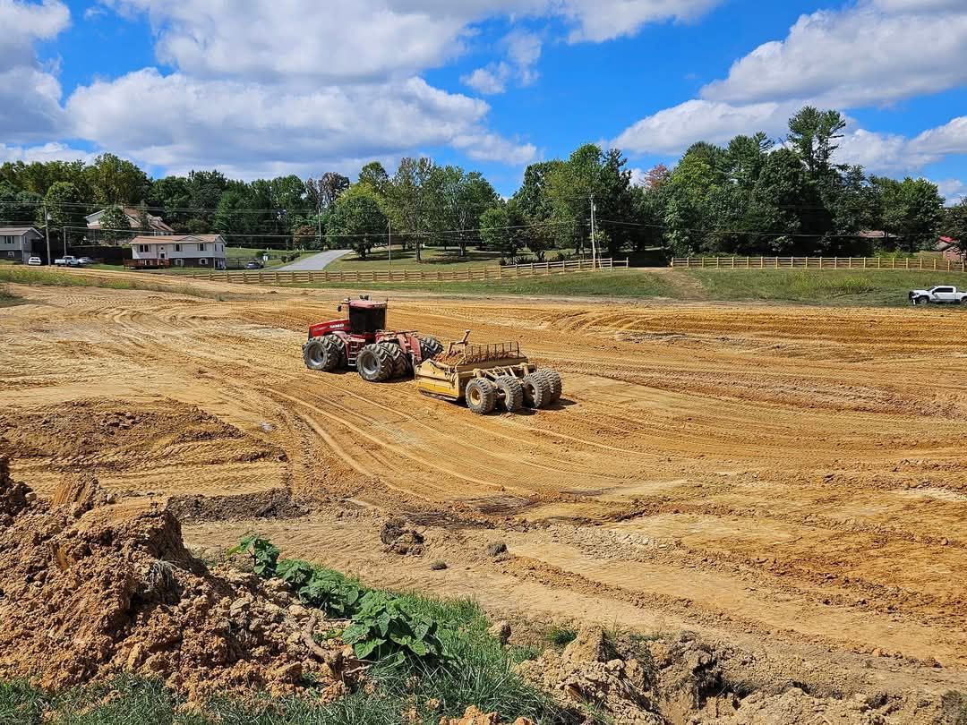 Tractor grading a dusty construction lot with earthmoving equipment under a blue sky