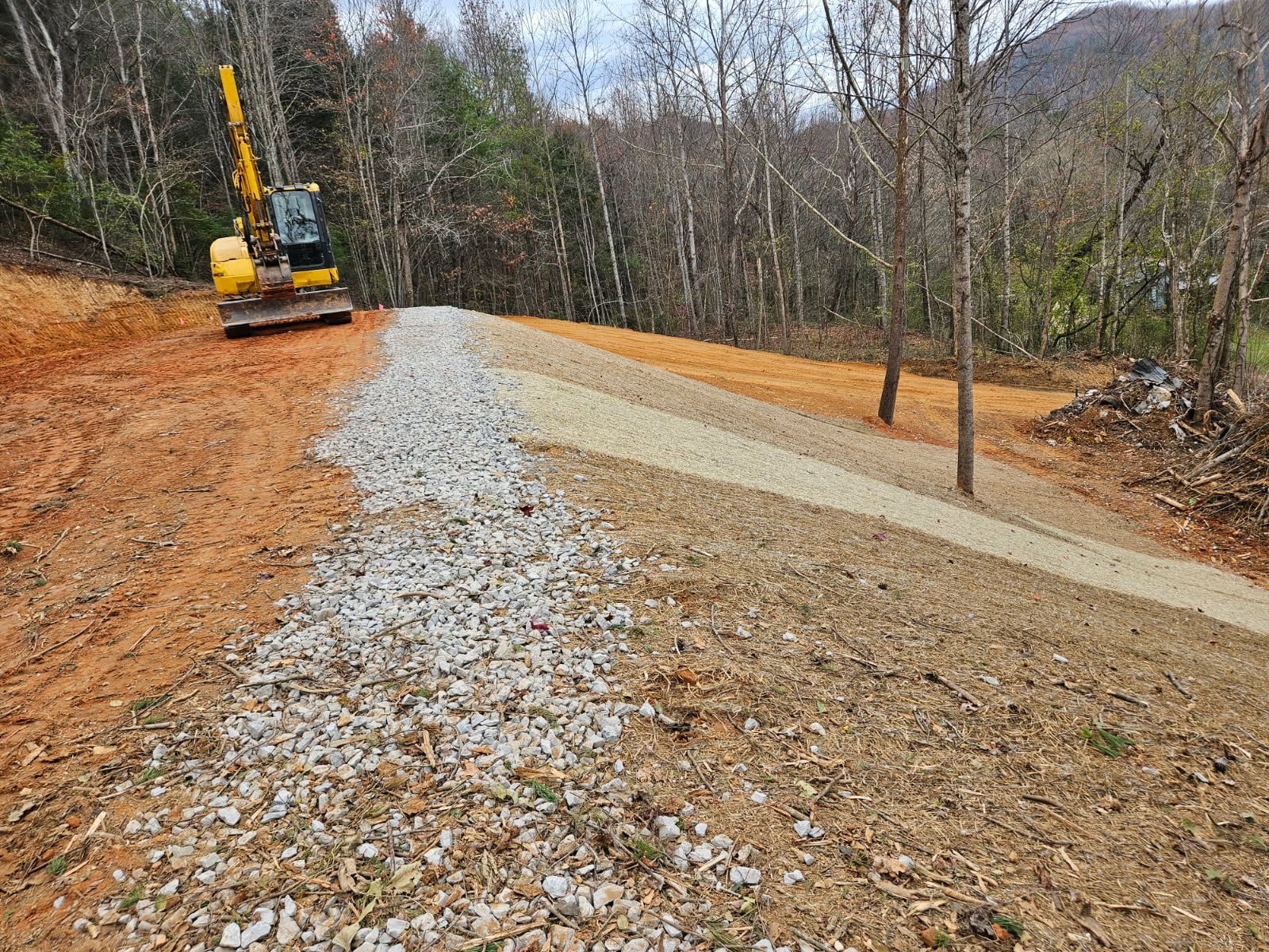 Gravel road under construction in a wooded hillside, with an excavator on the left and bare trees surrounding it