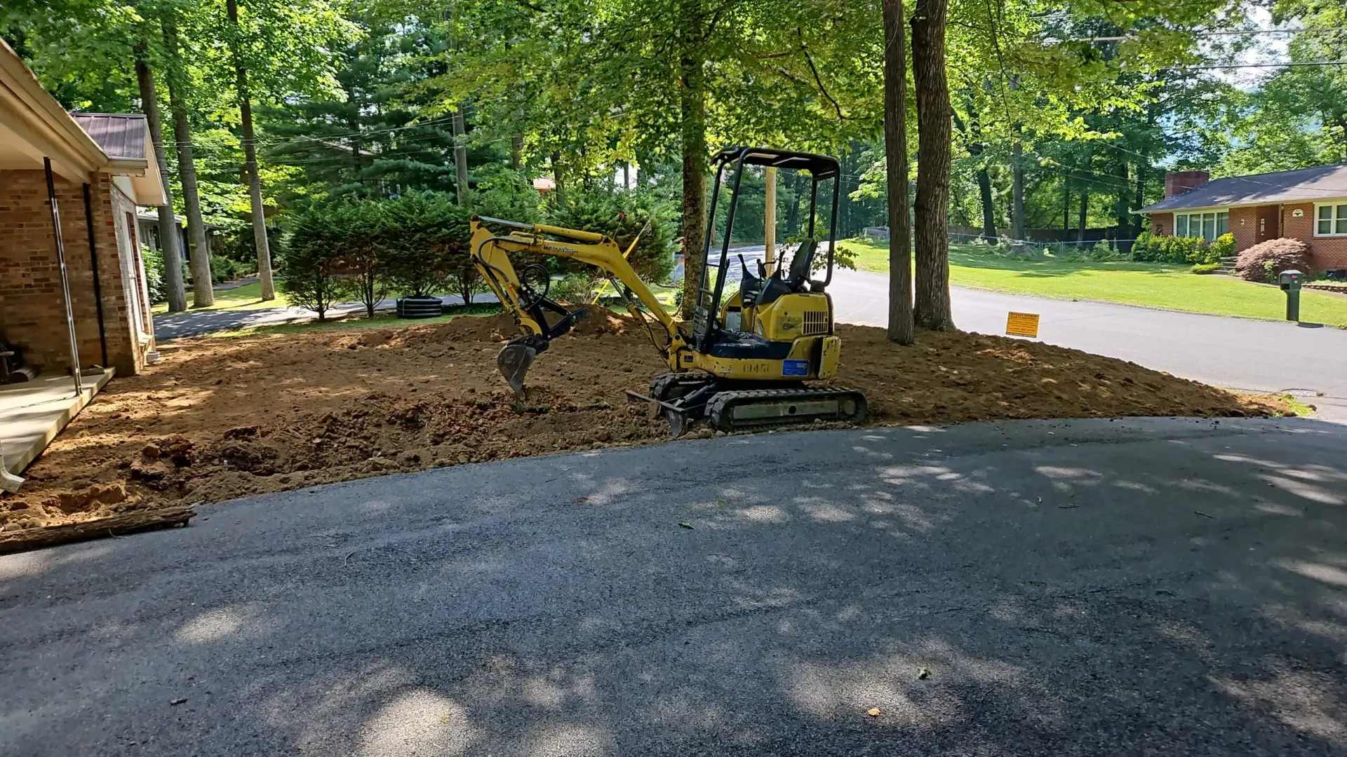 Yellow excavator clearing dirt beside a gravel driveway in a wooded residential area