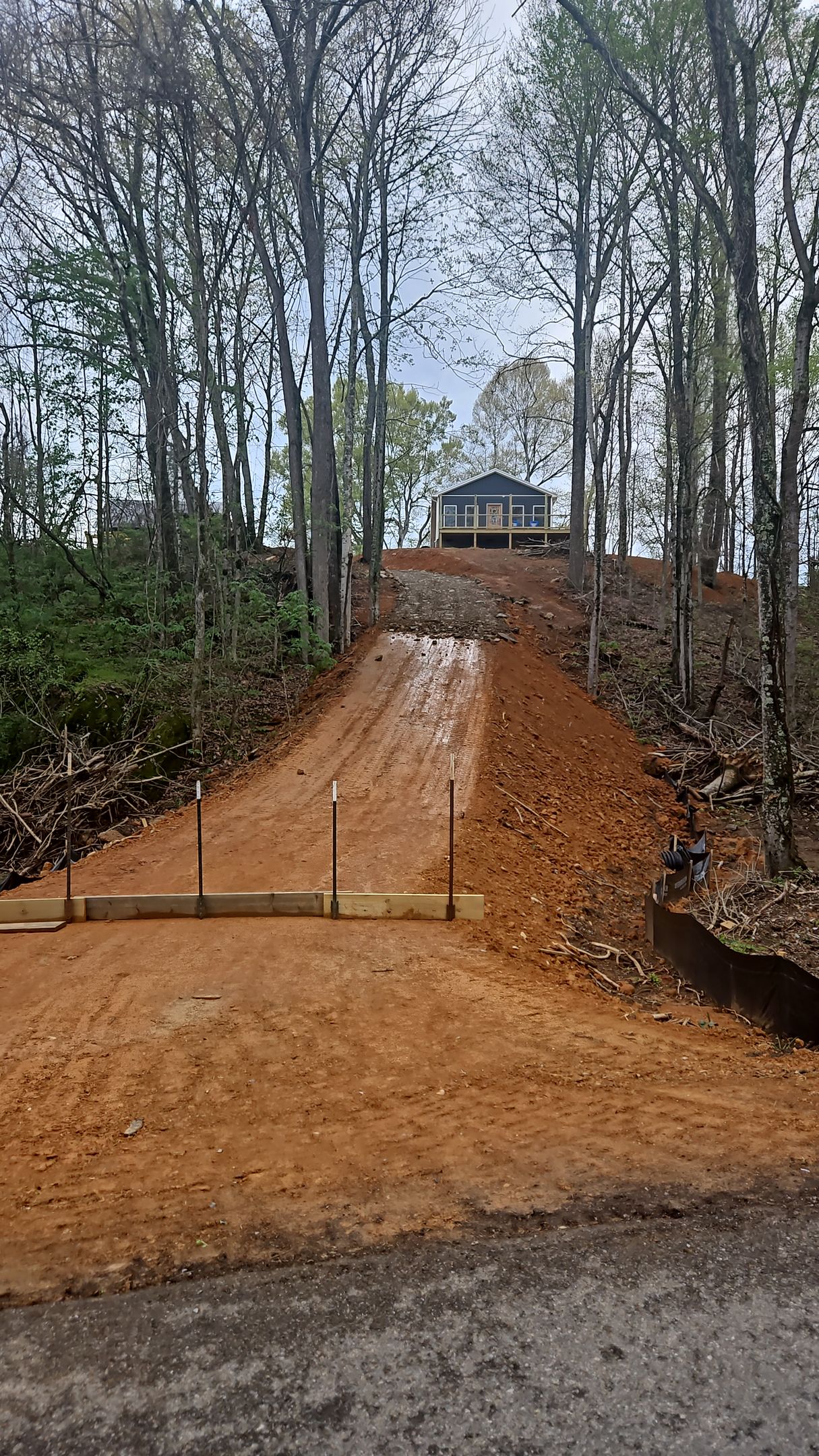 Steep dirt driveway leading to a house on a wooded hillside, with trees and overcast sky.
