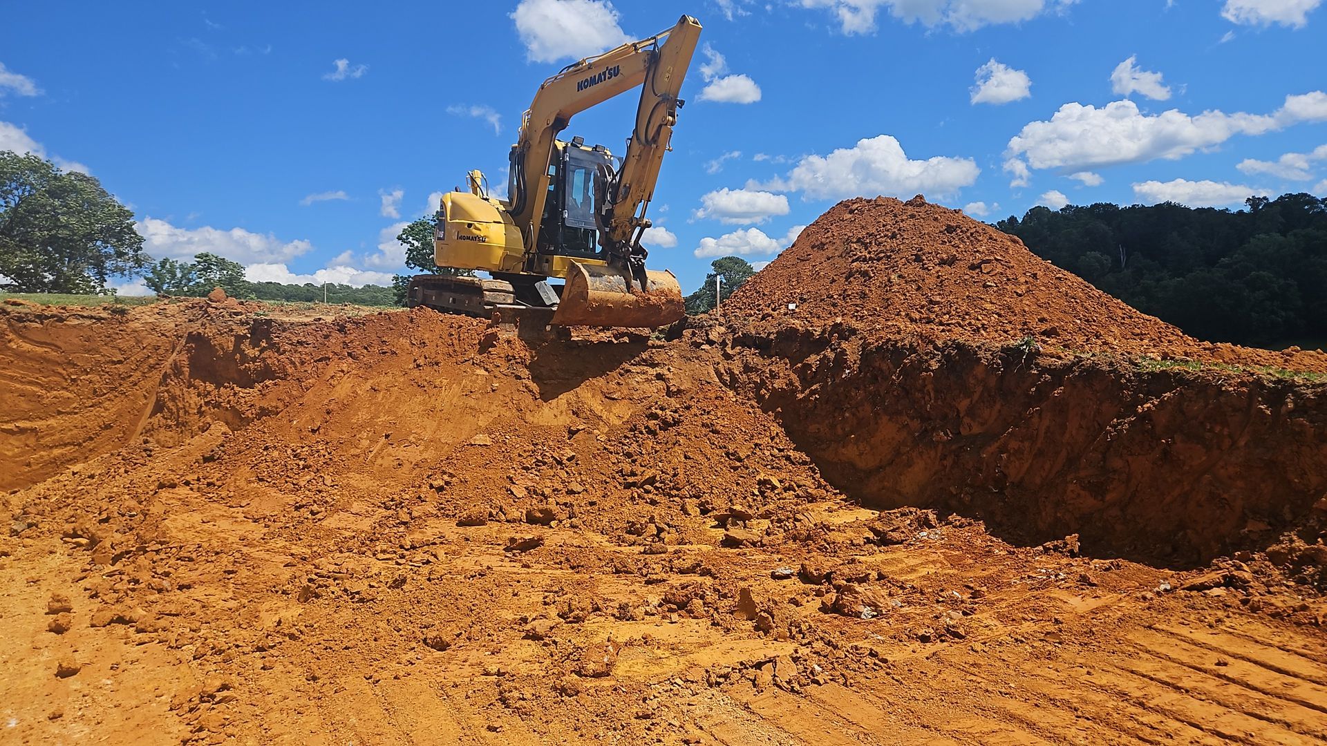 Yellow excavator on a dirt construction site beside a large soil pile under a blue sky.