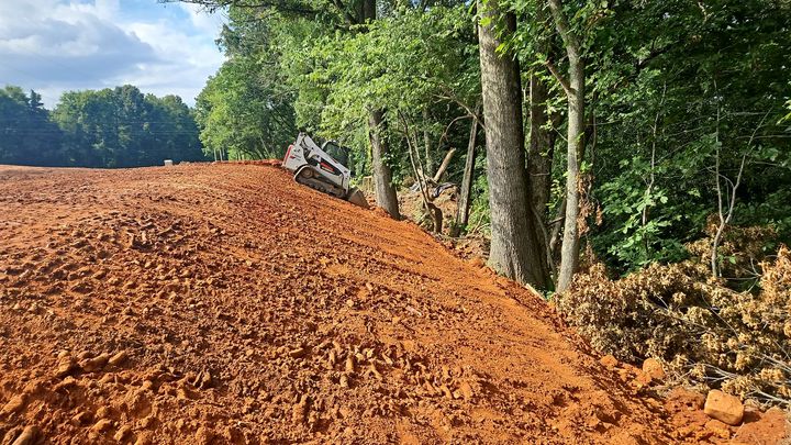 Red dirt road cut through trees, with a bulldozer working near the edge of a wooded area.