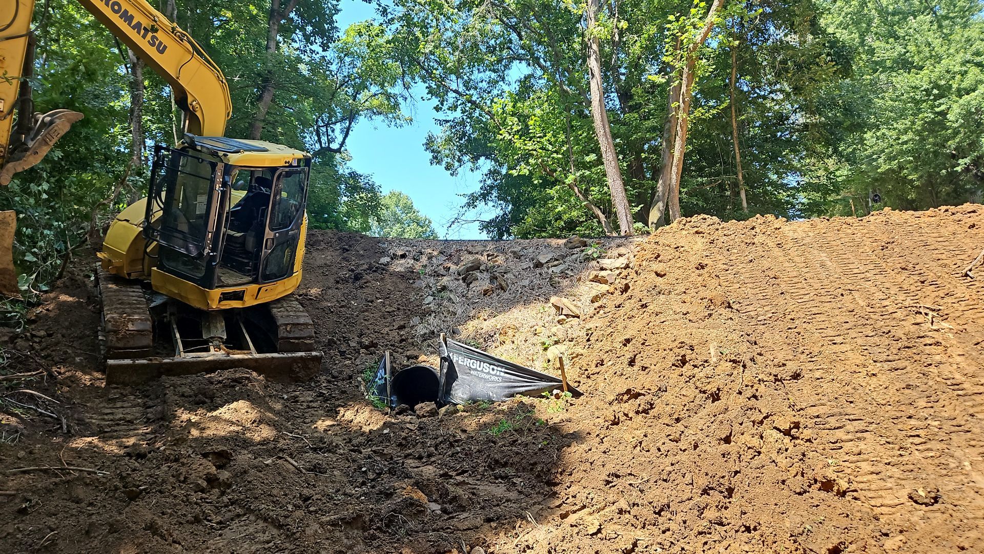Yellow excavator digging a trench beside a large dirt mound in a wooded area