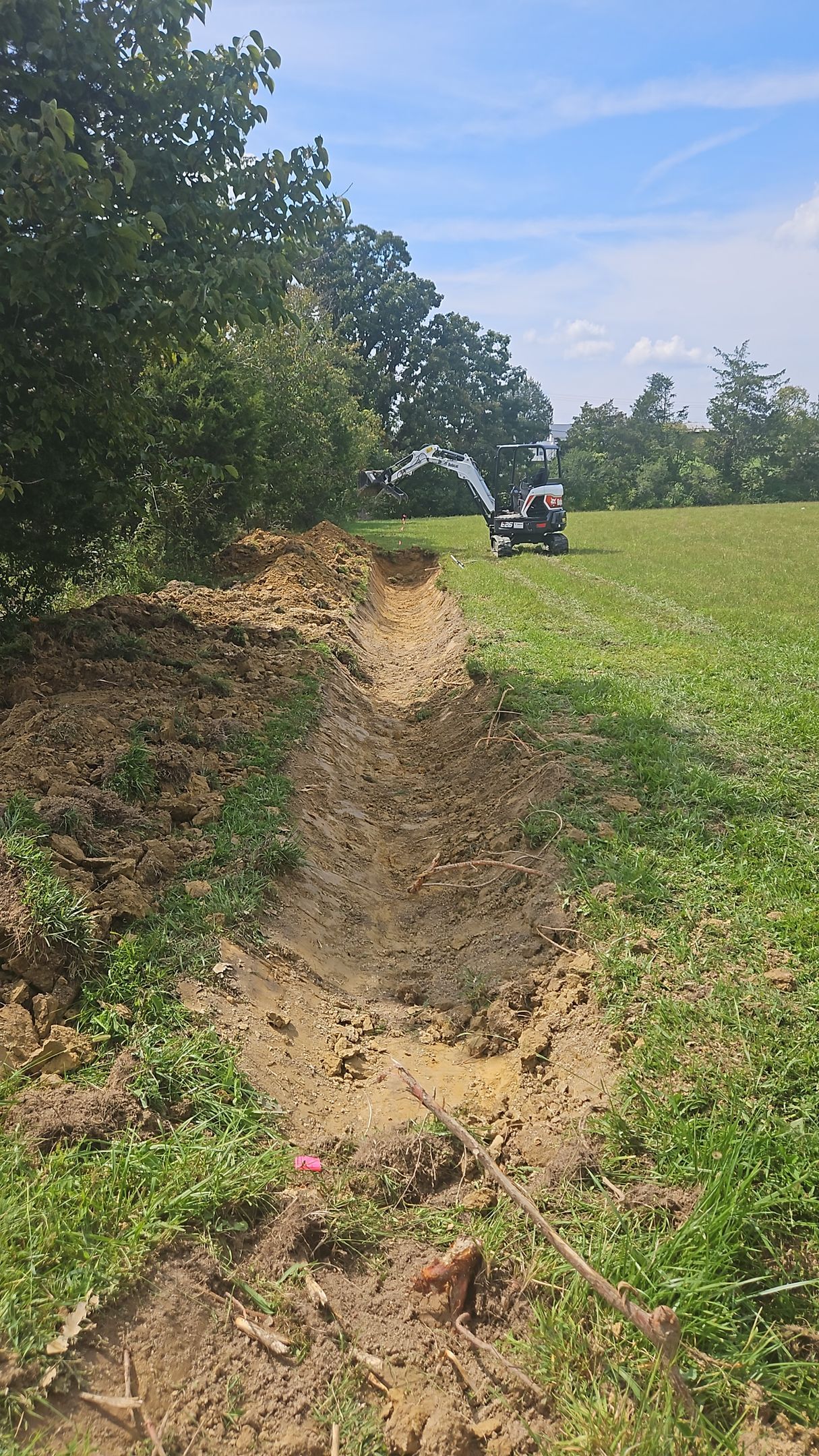 Narrow dirt trench through green field beside trees, with a small vehicle in the distance.
