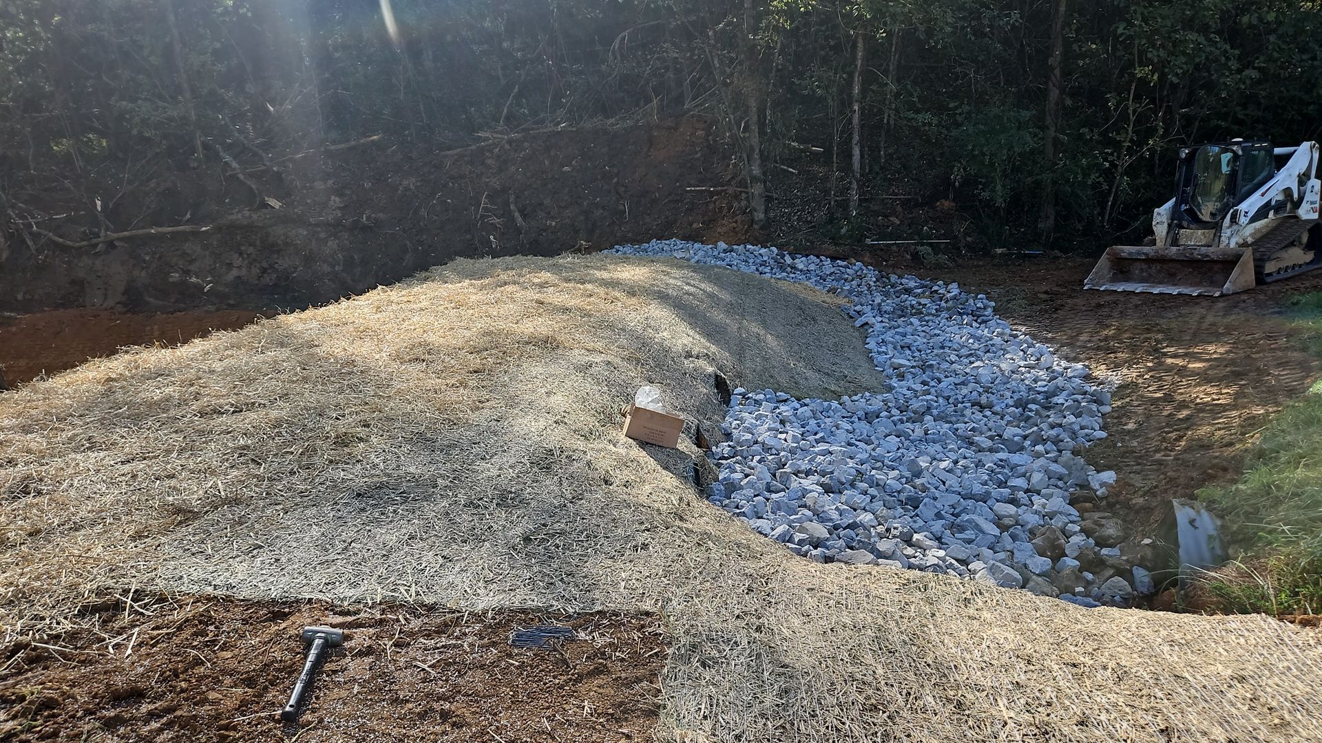 Gravel and dirt mound beside a blue-gray stone trench in a wooded yard, with an excavator in the background