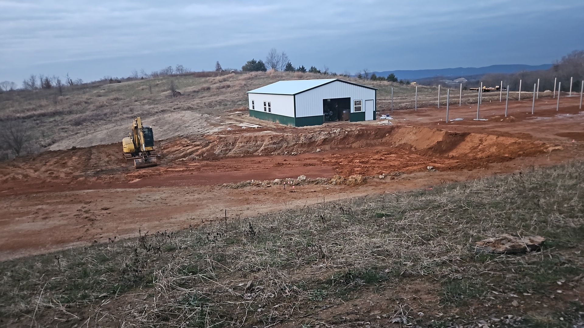Small white barn on a dirt hillside with a yellow excavator and overcast sky.
