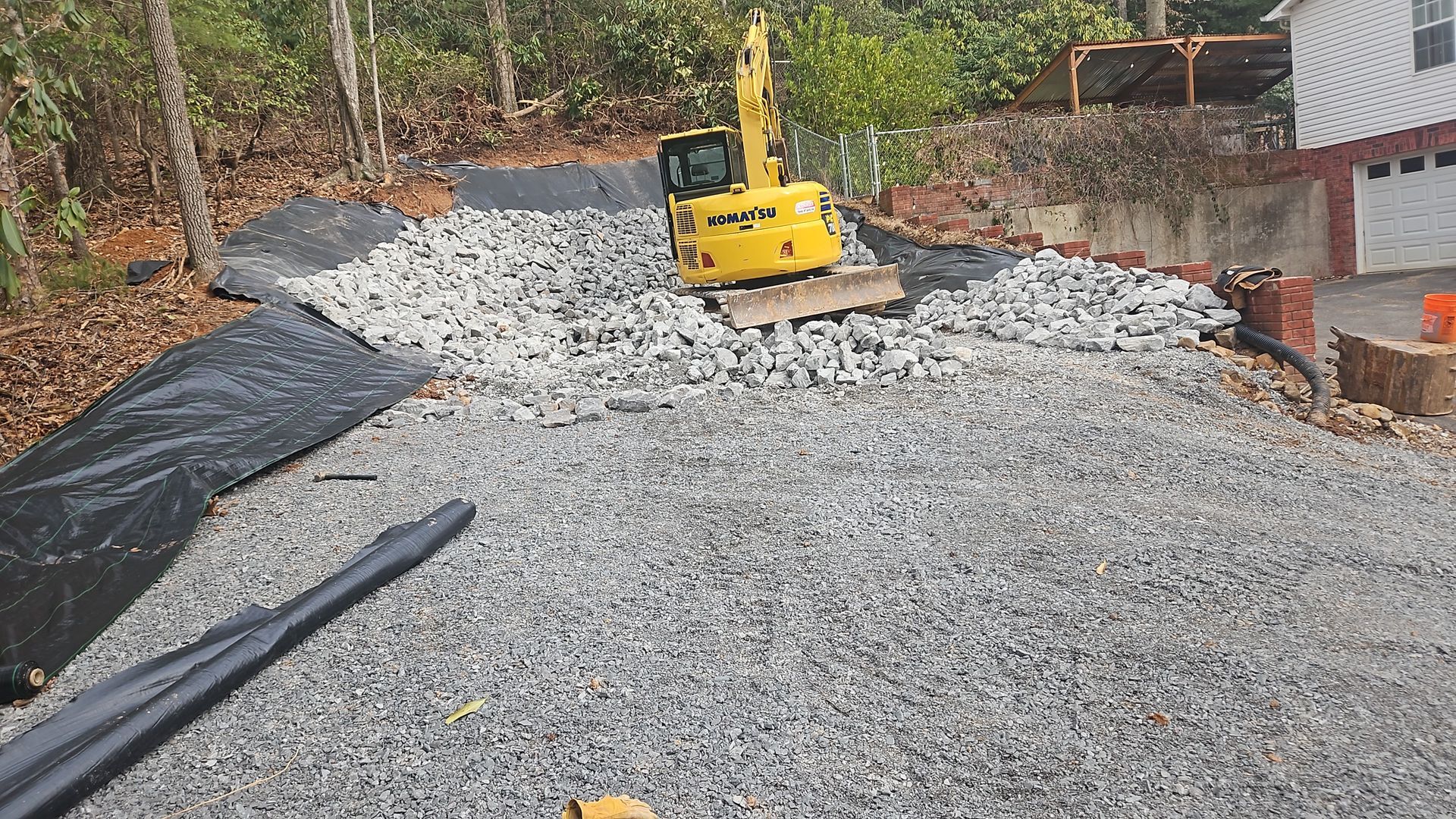 Yellow excavator on a gravel driveway under construction beside a house and wooded area