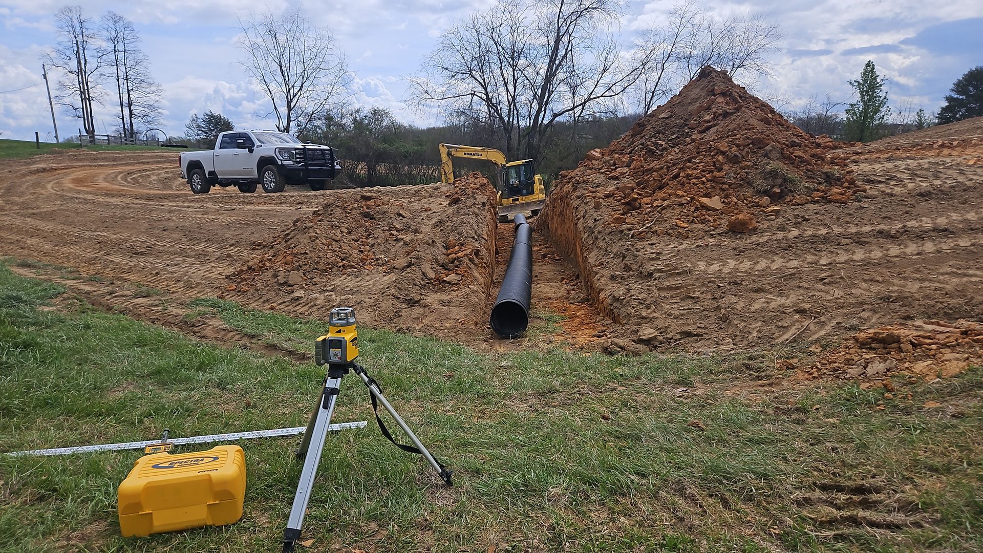 Construction site with white truck, yellow excavator, dirt pile, and large pipe on a bare lot