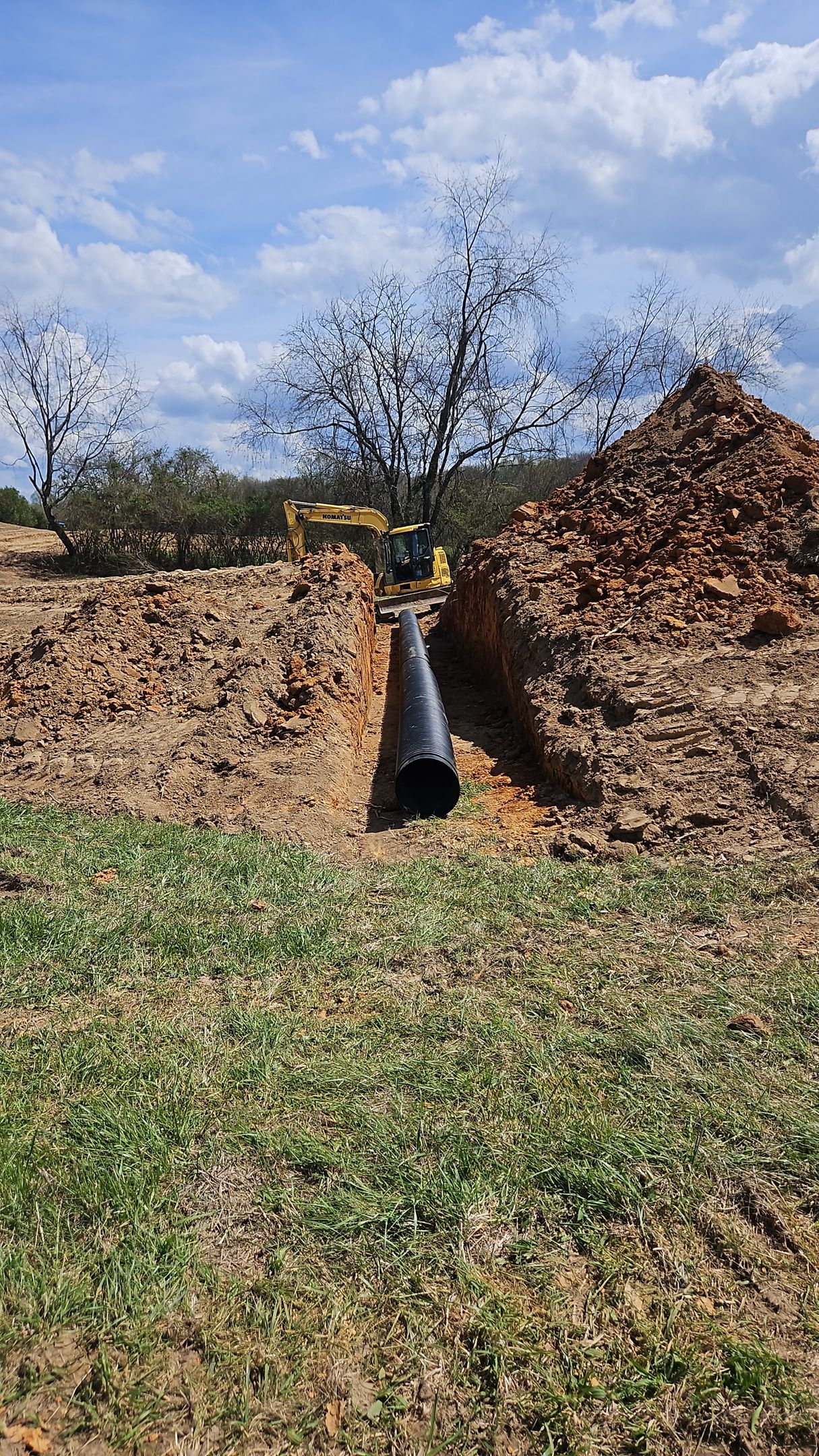 Excavator with bucket inside a muddy trench between dirt mounds under a blue sky