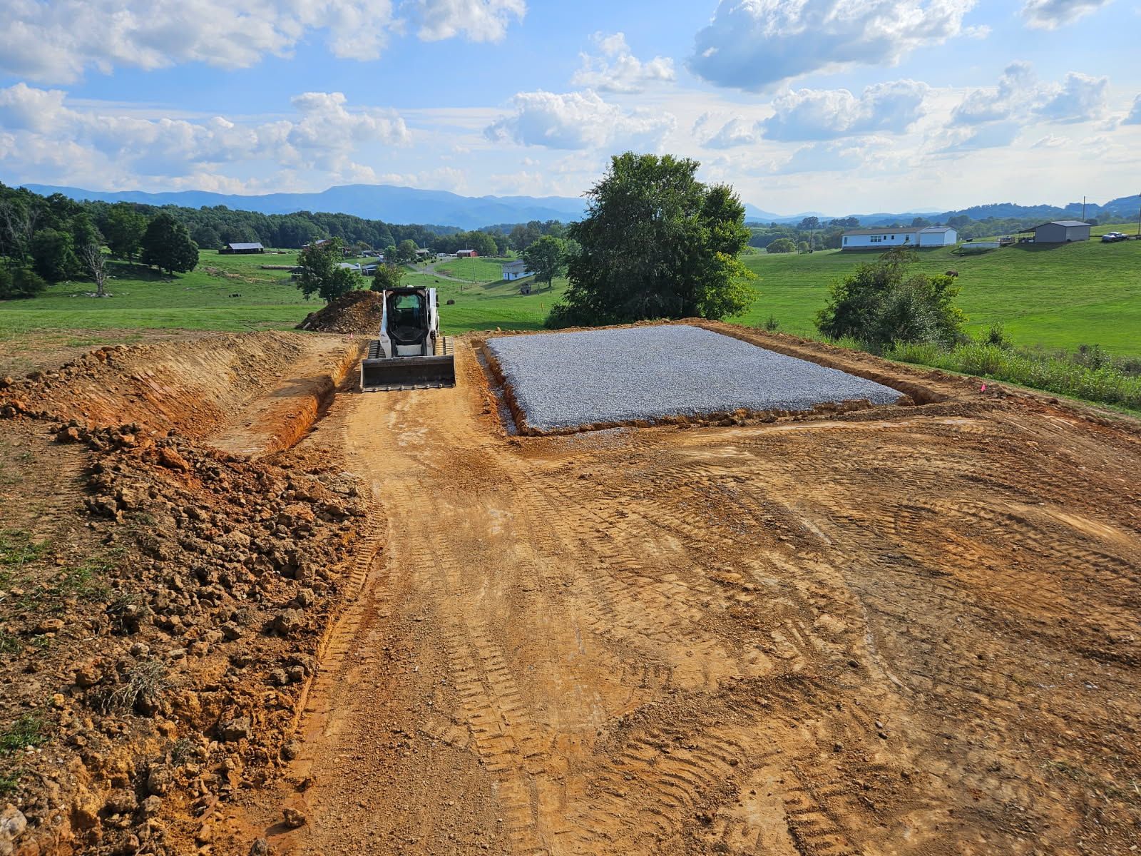 Bulldozer leveling a dirt road beside a gravel pad in a rural landscape under a cloudy sky
