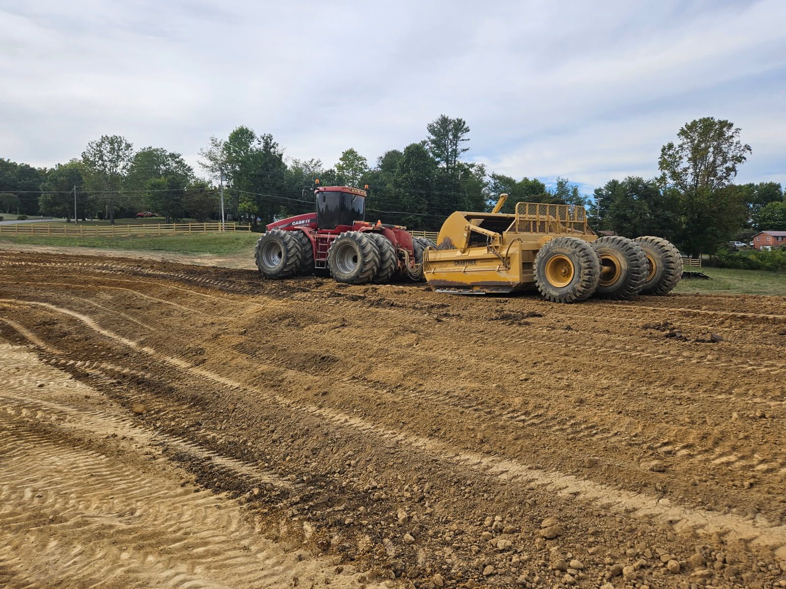 Red tractor and yellow bulldozer working on a muddy dirt field with trees in the background