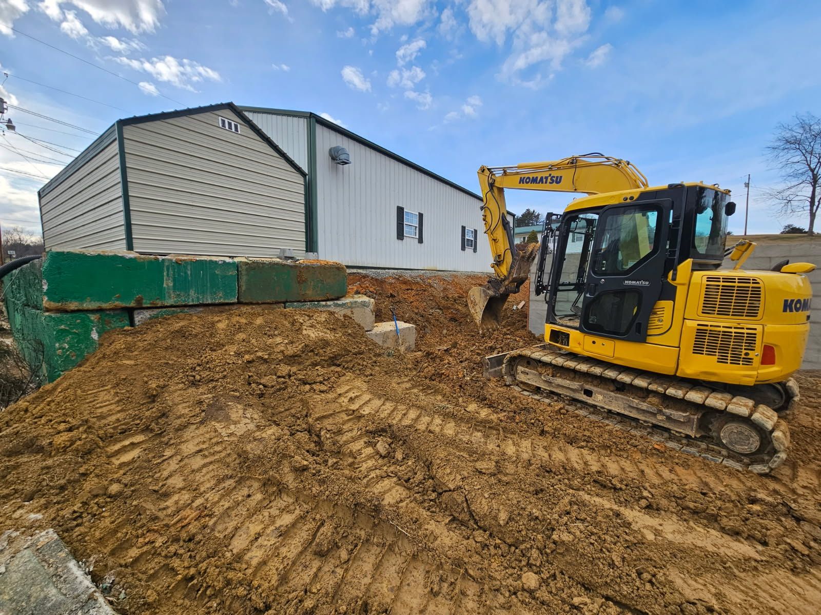 Yellow excavator moving dirt beside a green building under a blue sky