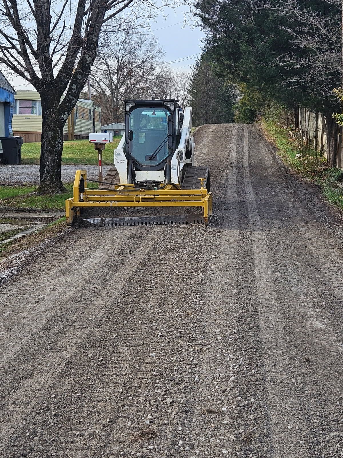 Compact tractor grading a gravel driveway lined with trees and fencing