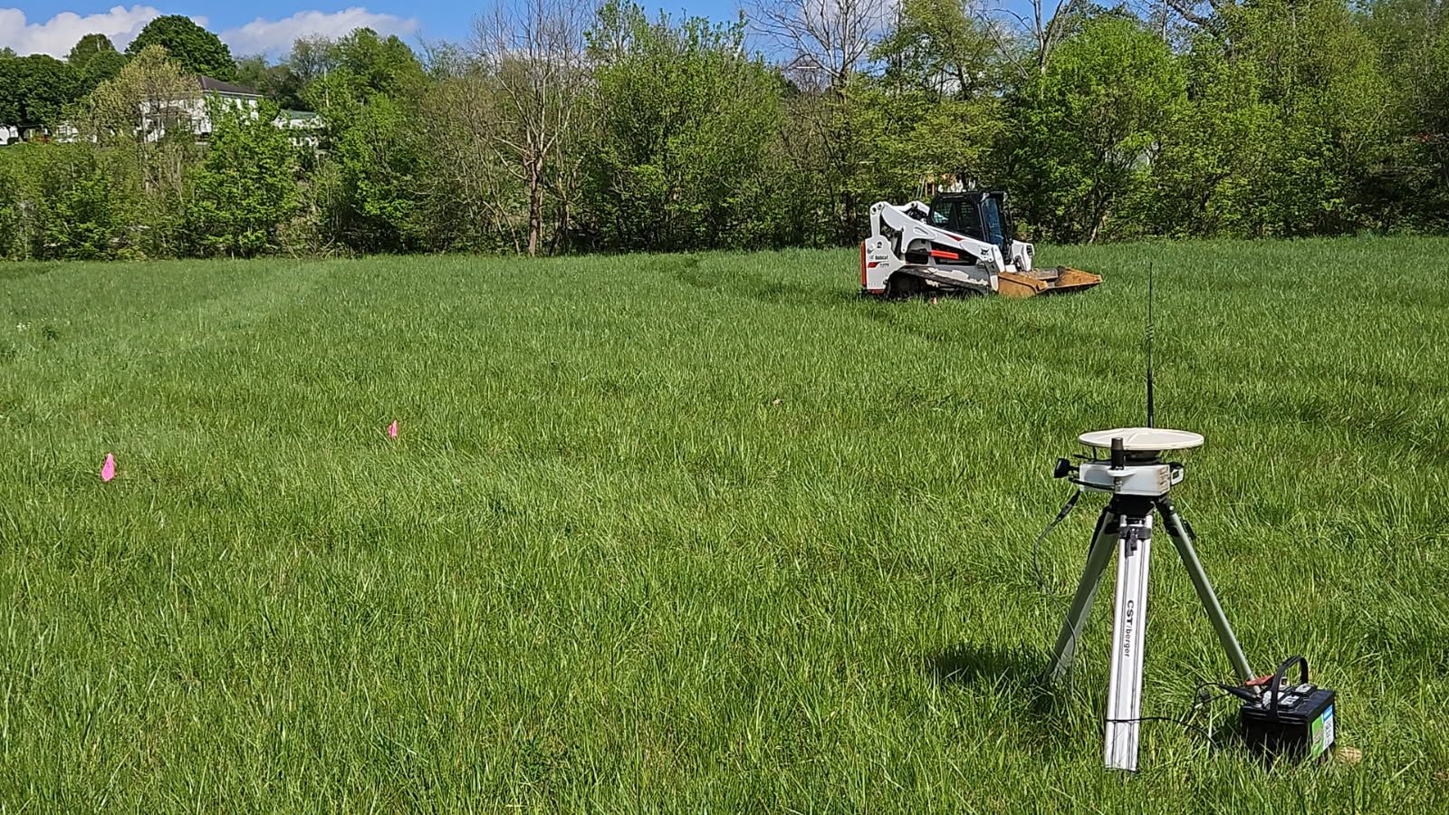 Surveying equipment in a green field with a small vehicle in the distance and trees along the edge