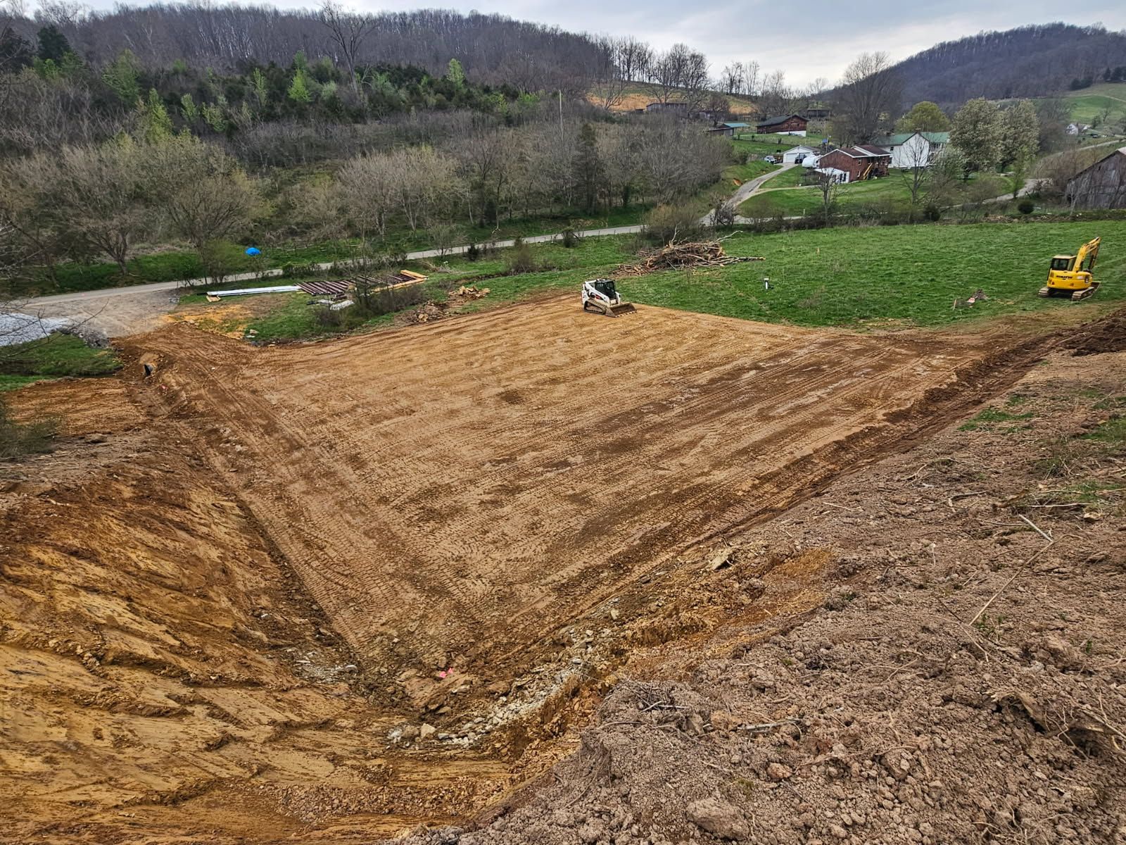 Excavated dirt field with a small excavator, surrounded by green hills and bare trees under cloudy skies