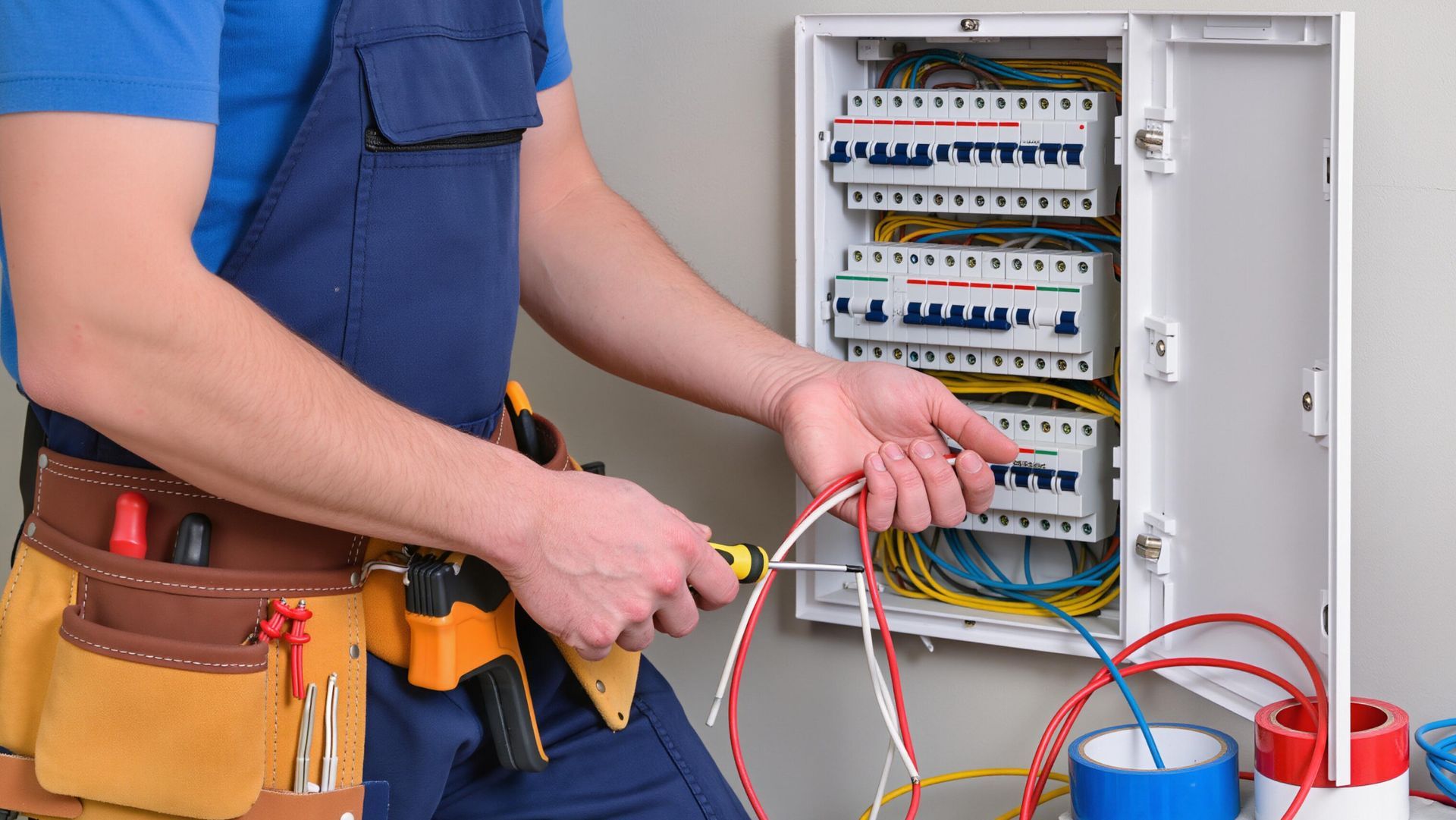 Un électricien effectue le câblage d'un tableau électrique ouvert avec des fils rouges et bleus, portant une ceinture à outils.