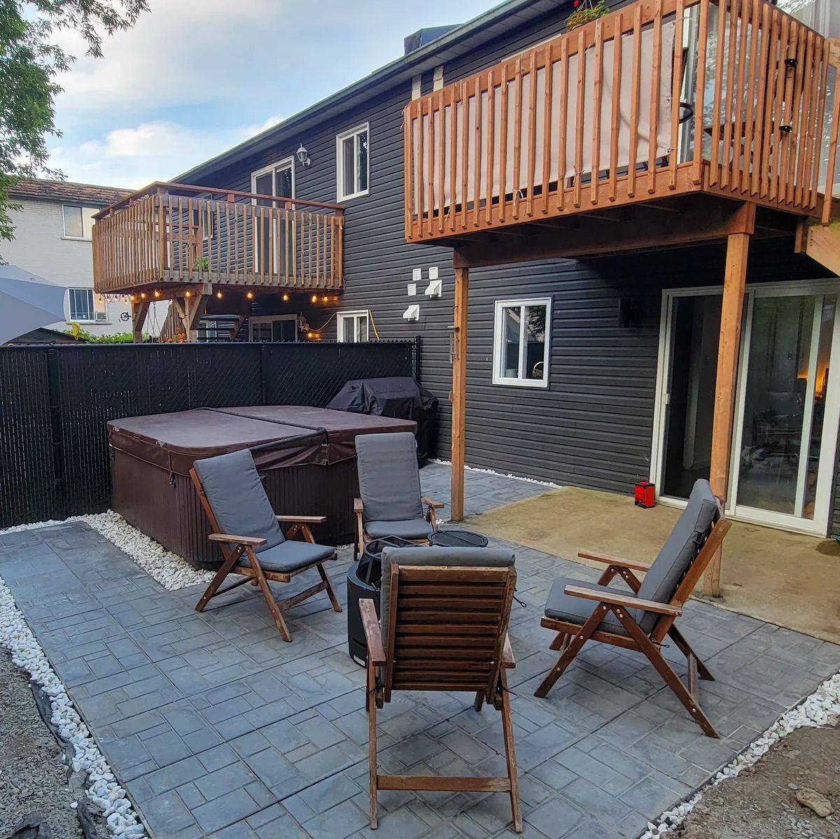 Terrasse arrière avec dalles grises, chaises pliantes marron, jacuzzi et deux balcons en bois sur une maison sombre
