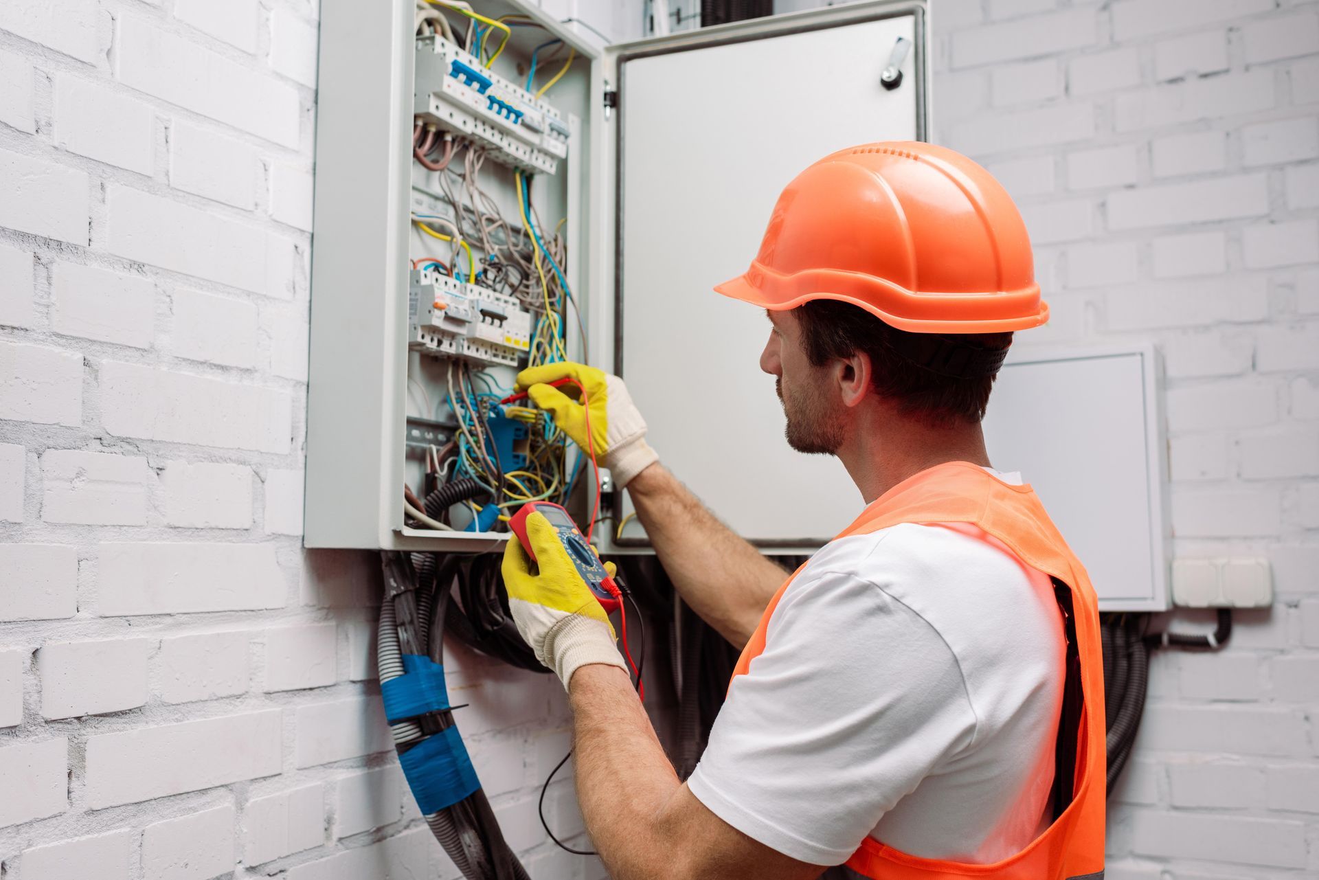 Électricien portant un casque et un gilet orange, effectuant le câblage d'un tableau électrique ouvert à l'intérieur.