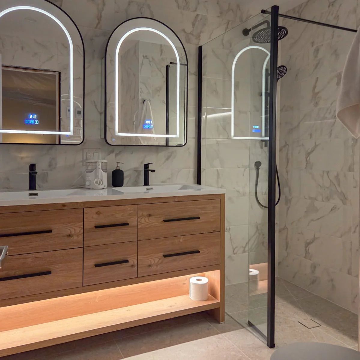 Salle de bains moderne avec meuble-lavabo en bois, miroirs arqués, murs en pierre blanche et cabine de douche en verre.