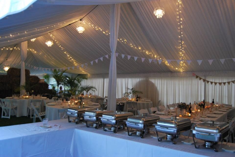 Buffet tables with chafing dishes set up in a draped, white-lit tent for a formal event with guest seating in the background.