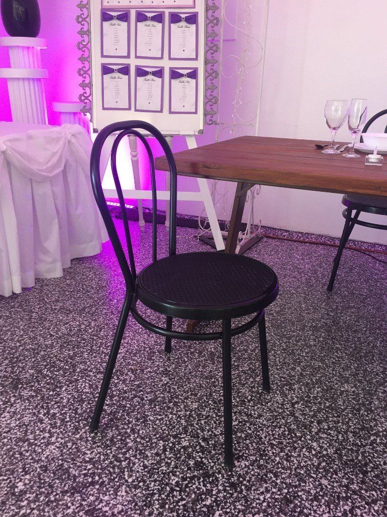 A black metal chair sits on a speckled floor in front of a rustic wooden table and a white seating chart display.