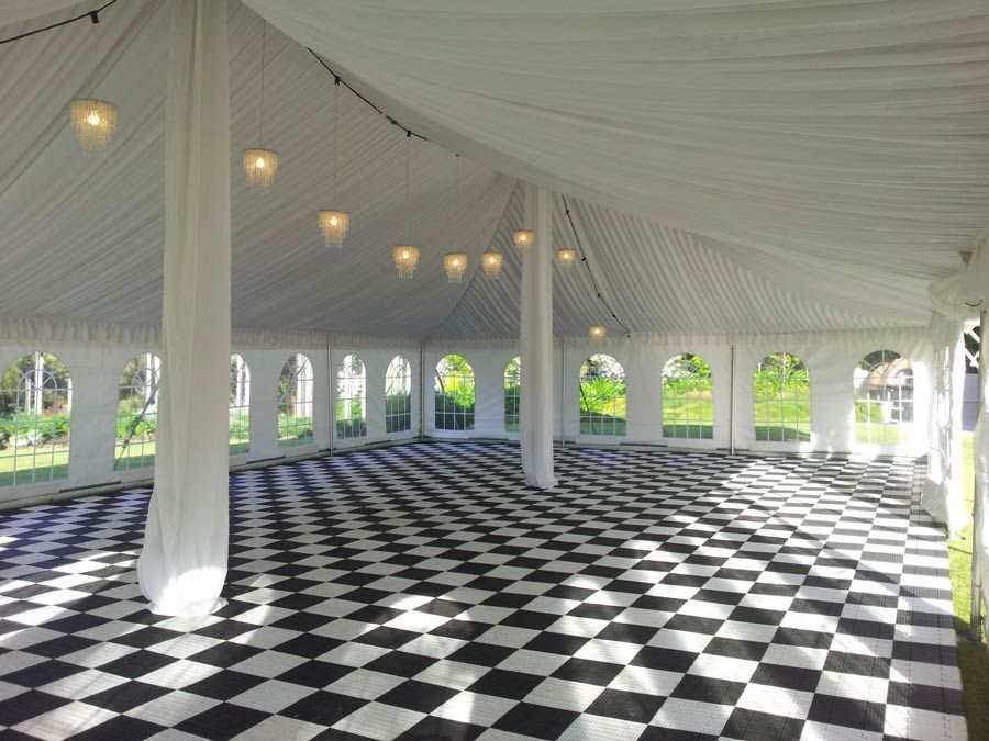 A white tent interior with a black-and-white checkered floor, arched windows, and string lights hanging from the ceiling.