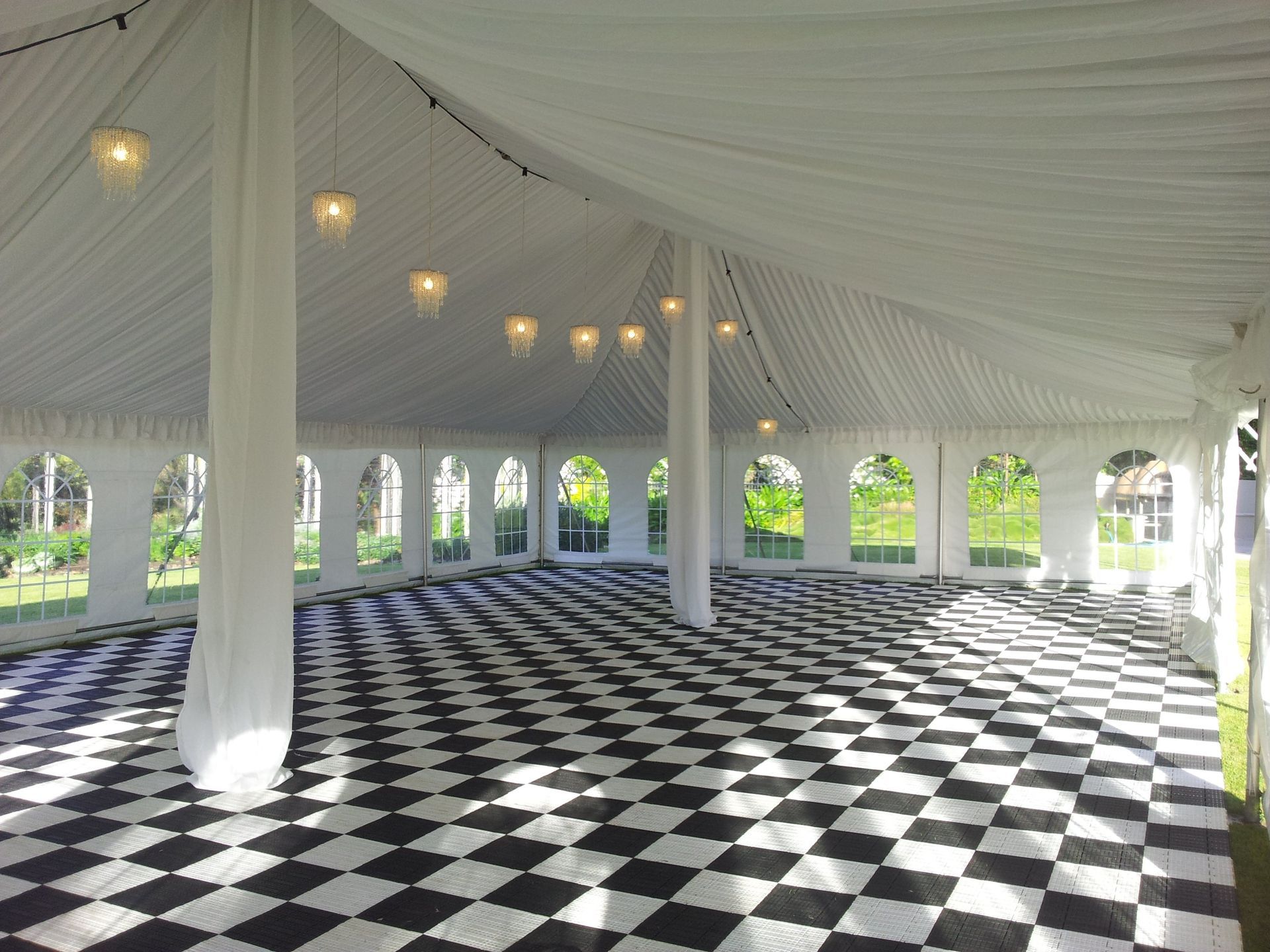 An empty white tent featuring a black-and-white checkered dance floor, draped fabric ceilings, and hanging lights.