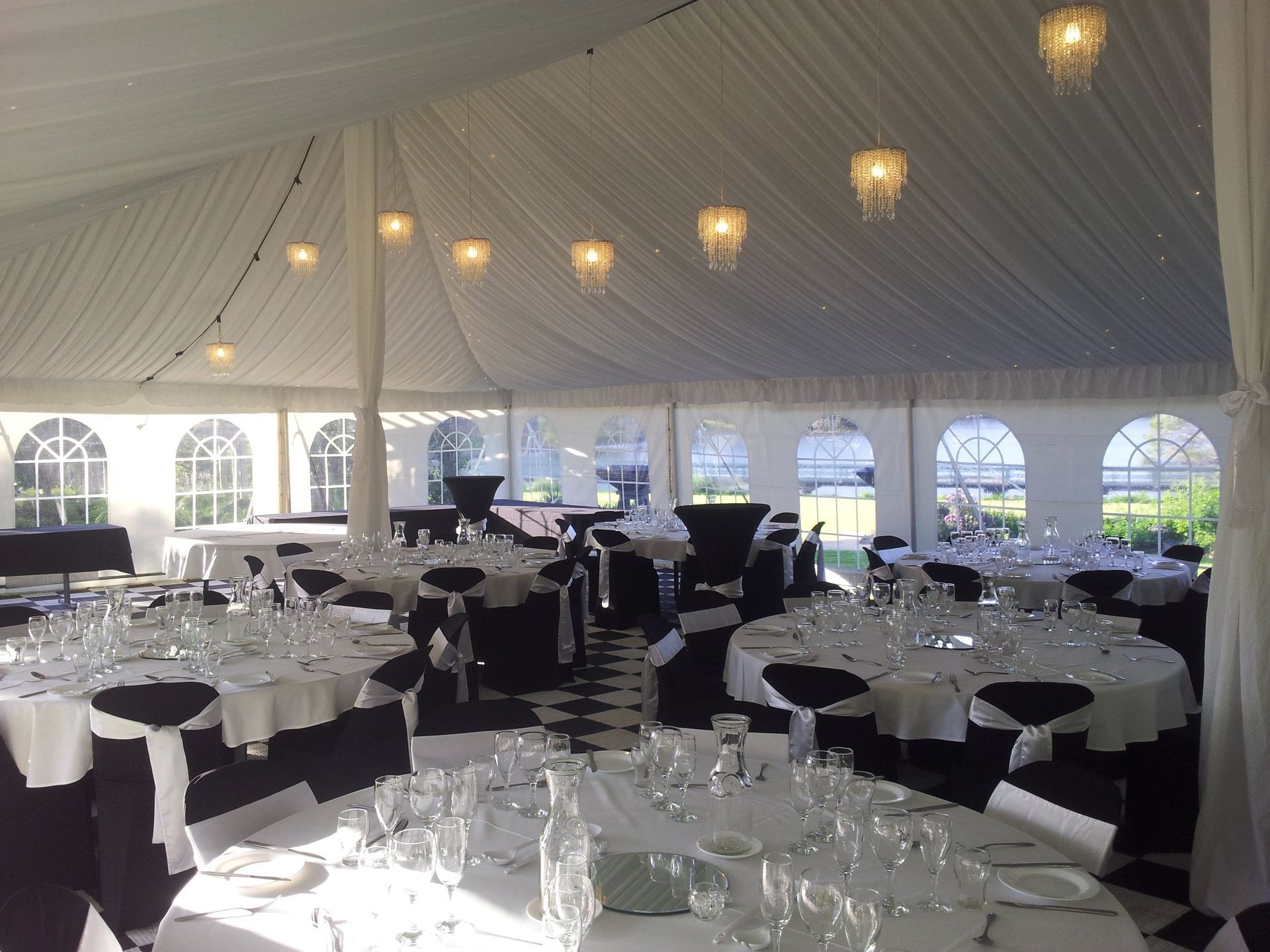 A bright wedding reception tent featuring round tables with white linens, black chair covers, and hanging globe lights.