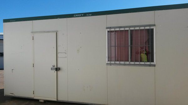 A portable, off-white modular office building with a single door and a barred window against a clear blue sky.