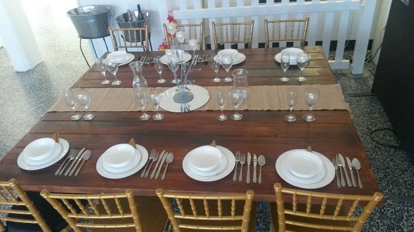 A rustic wooden table set for dinner with white plates, glassware, and gold chairs in a brightly lit indoor space.