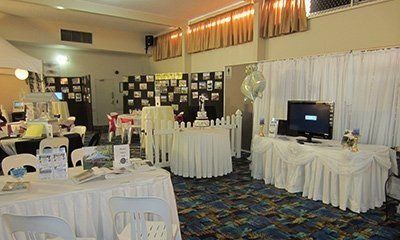 A banquet room set for a memorial or celebration with tables covered in white linens, photo boards, and a TV display.