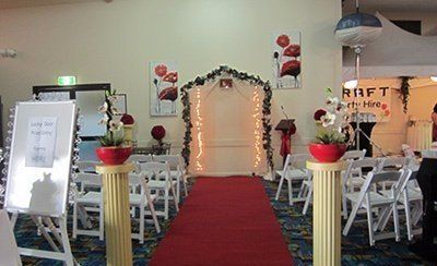 An indoor wedding ceremony setup featuring a red carpet leading to an arched, illuminated structure with floral decor.