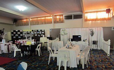 An event hall with white-clothed tables, chairs, decorations, and display boards against a wall under warm lighting.