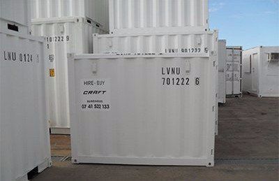 White shipping containers stacked and standing in an outdoor storage yard under a blue sky.
