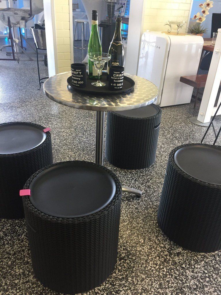 A high-top table with two wine bottles and a glass sits surrounded by four black wicker-style stools on a speckled floor.