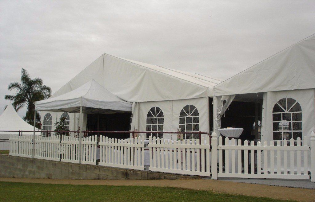 Large white event tents with arched windows and a white picket fence, set against a cloudy sky over a grassy lawn.